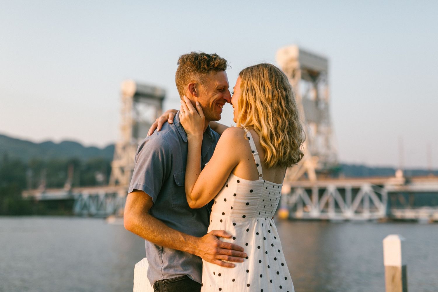 Intimate moment between couple on waterfront dock during sunset with bridge in background.