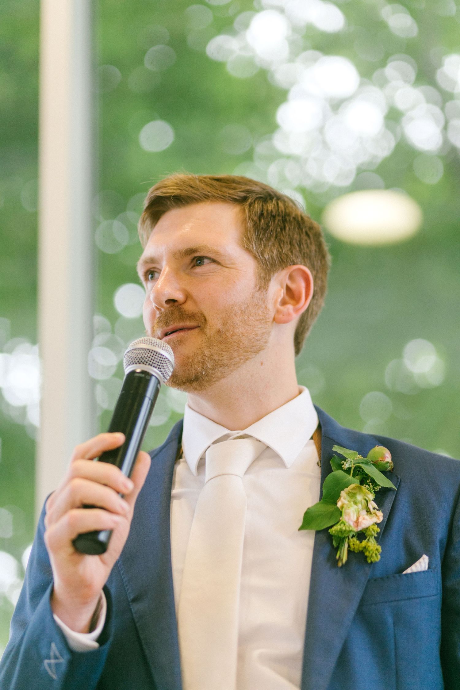 A speaker in a navy suit and light tie delivers a speech outdoors with a floral boutonniere.