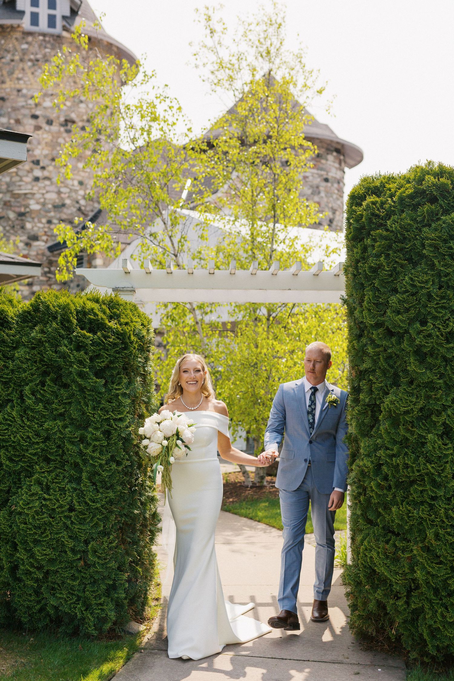 Wedding couple walking through a garden path lined with manicured hedges and trees.