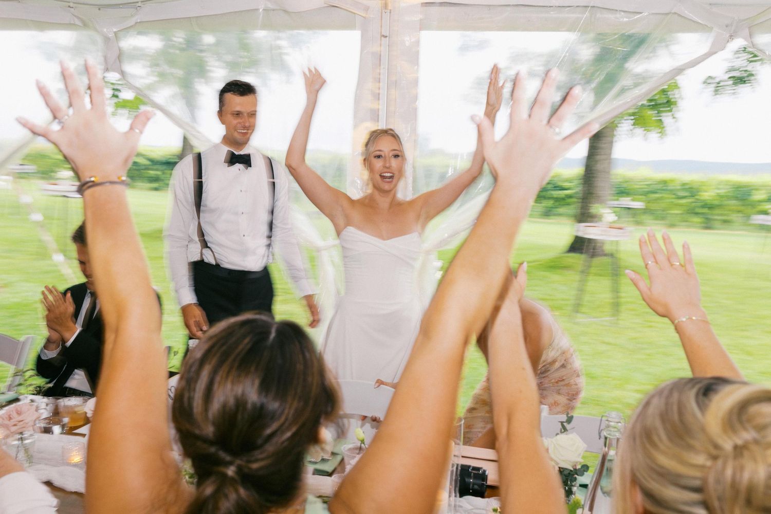 Wedding guests raise their hands in celebration under an outdoor tent during a joyful reception party.