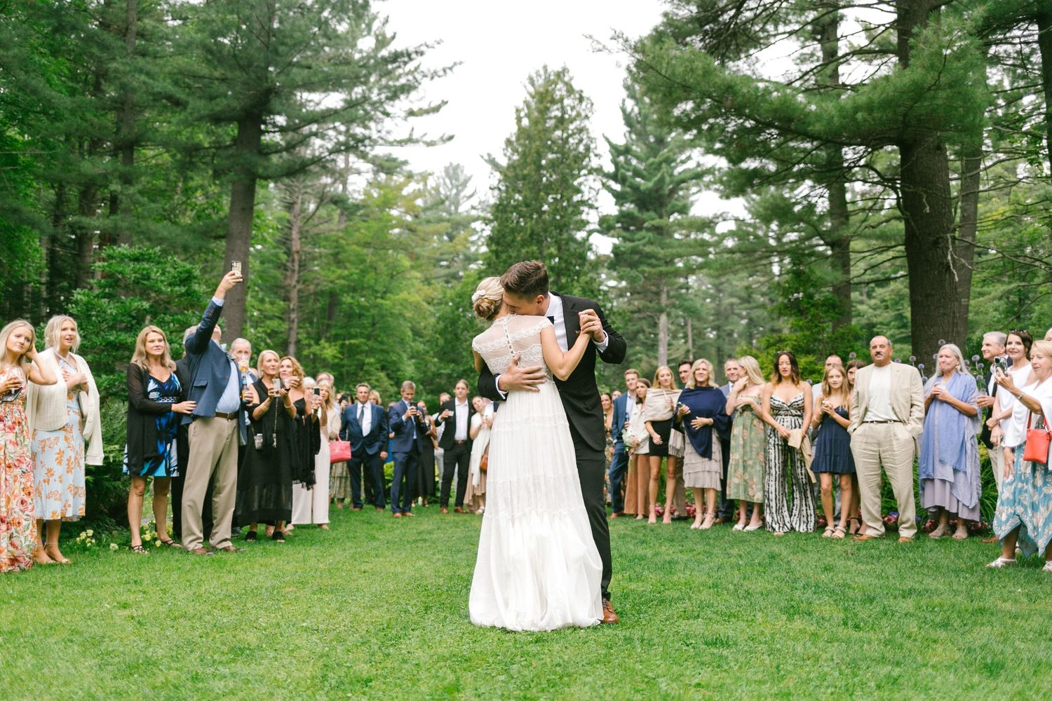 Wedding guests gather on a lush green lawn to watch the bride and groom's first dance surrounded by tall pine trees in a garden environment.