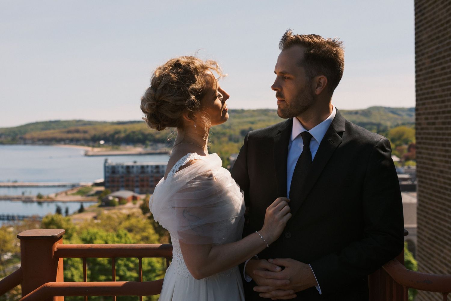A couple in wedding attire embraces on a balcony overlooking a scenic harbor and green hills on a sunny day.