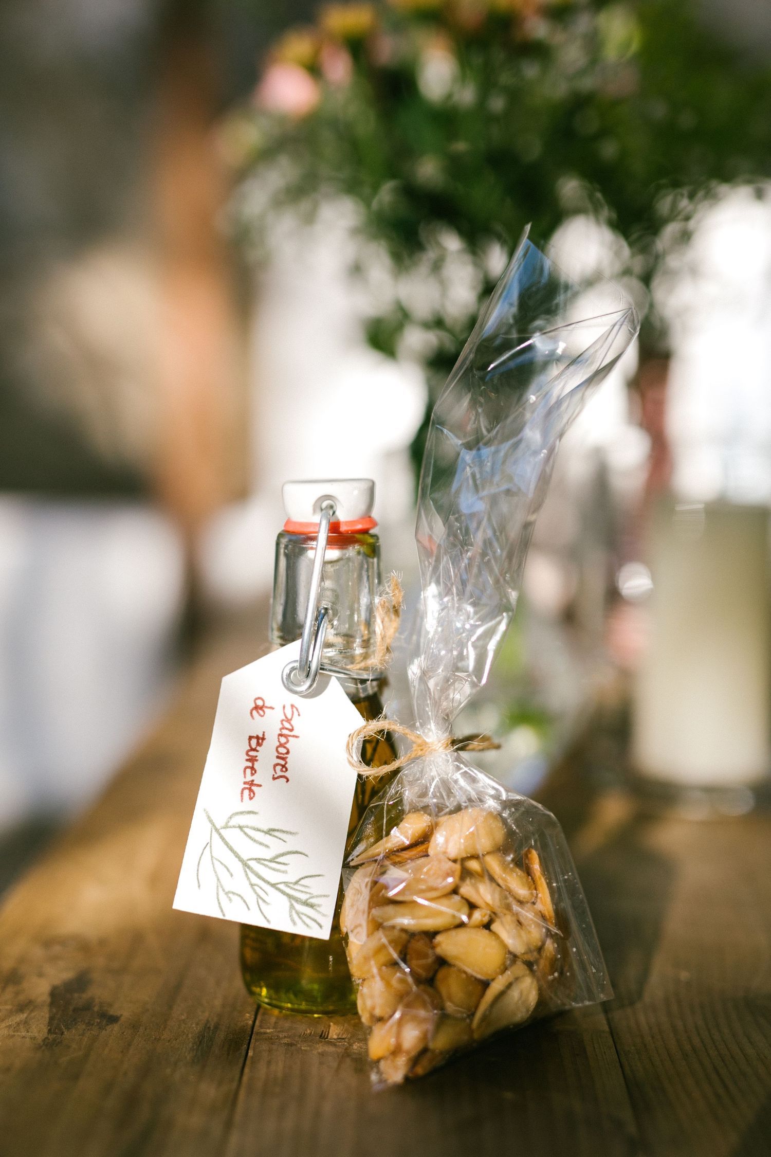 Small glass bottle of olive oil with handwritten tag next to clear bag of almonds on rustic wooden surface.