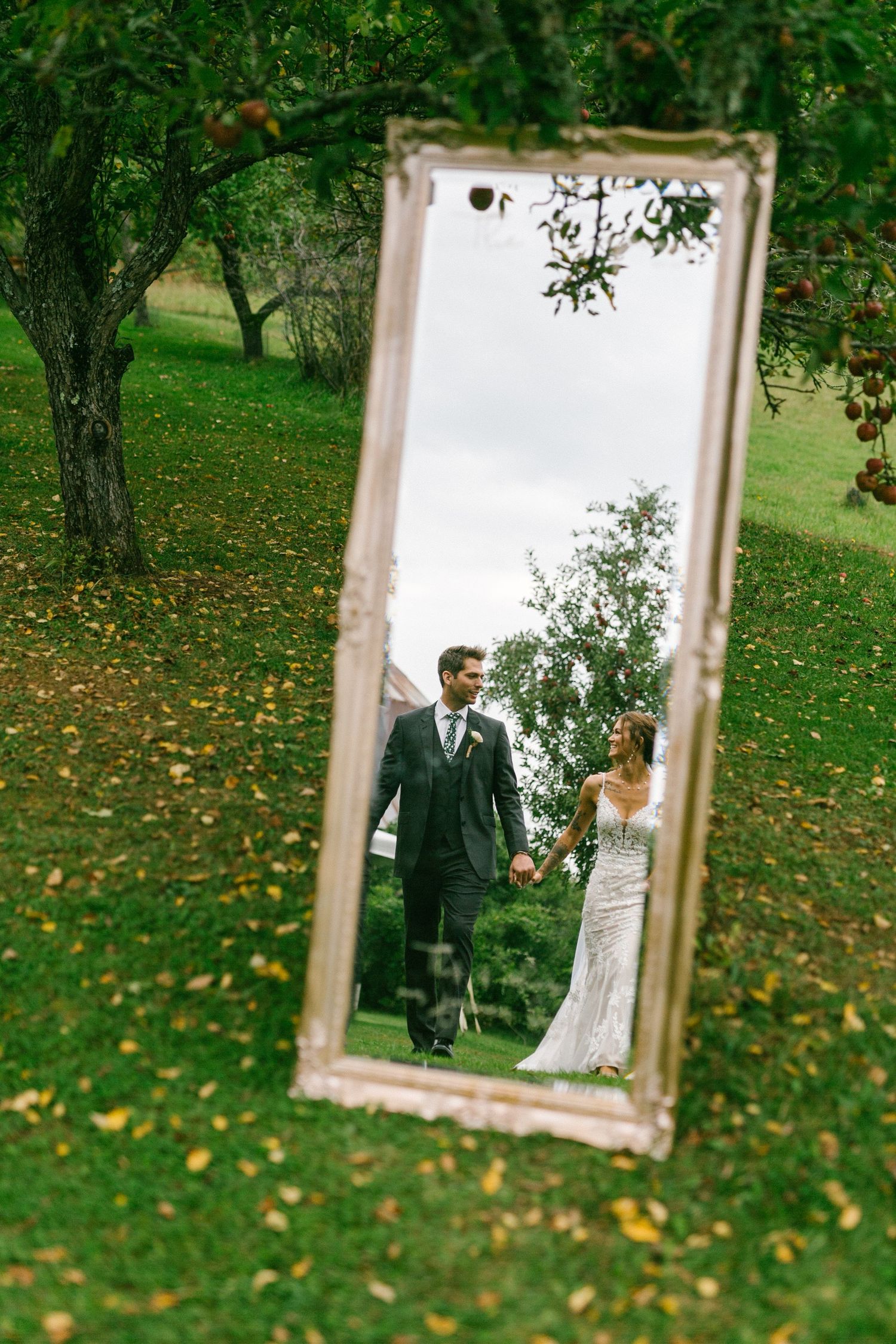 A vintage mirror placed on grass reflects a romantic wedding couple in an apple orchard with fallen autumn leaves.