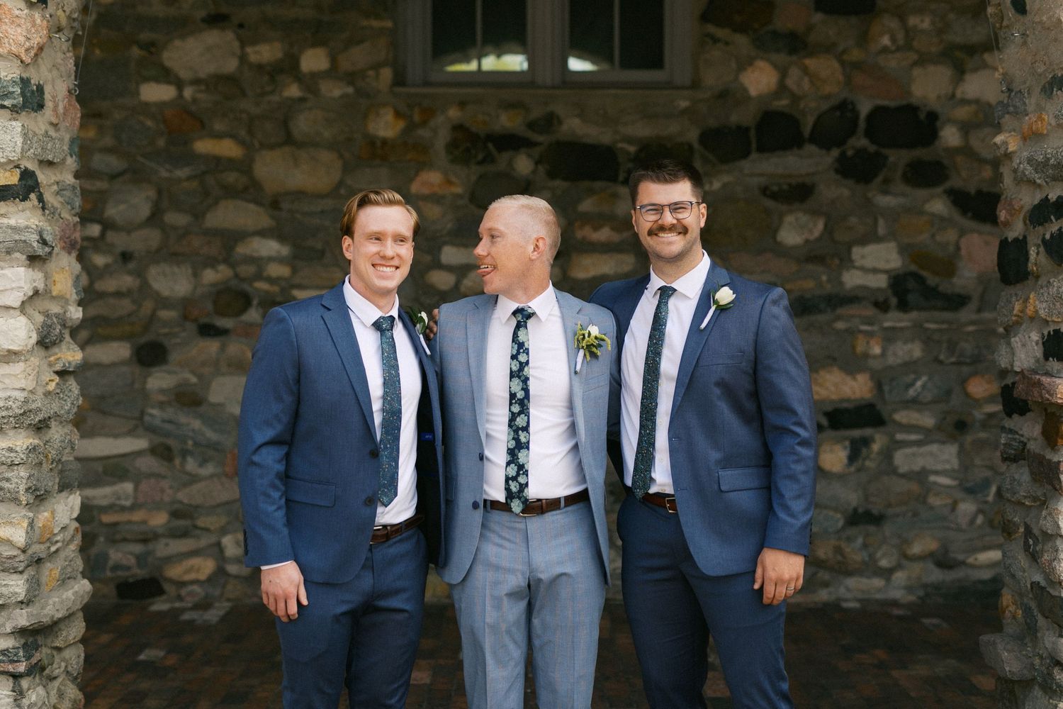 Wedding party members in navy and grey suits stand by a stone wall exterior.