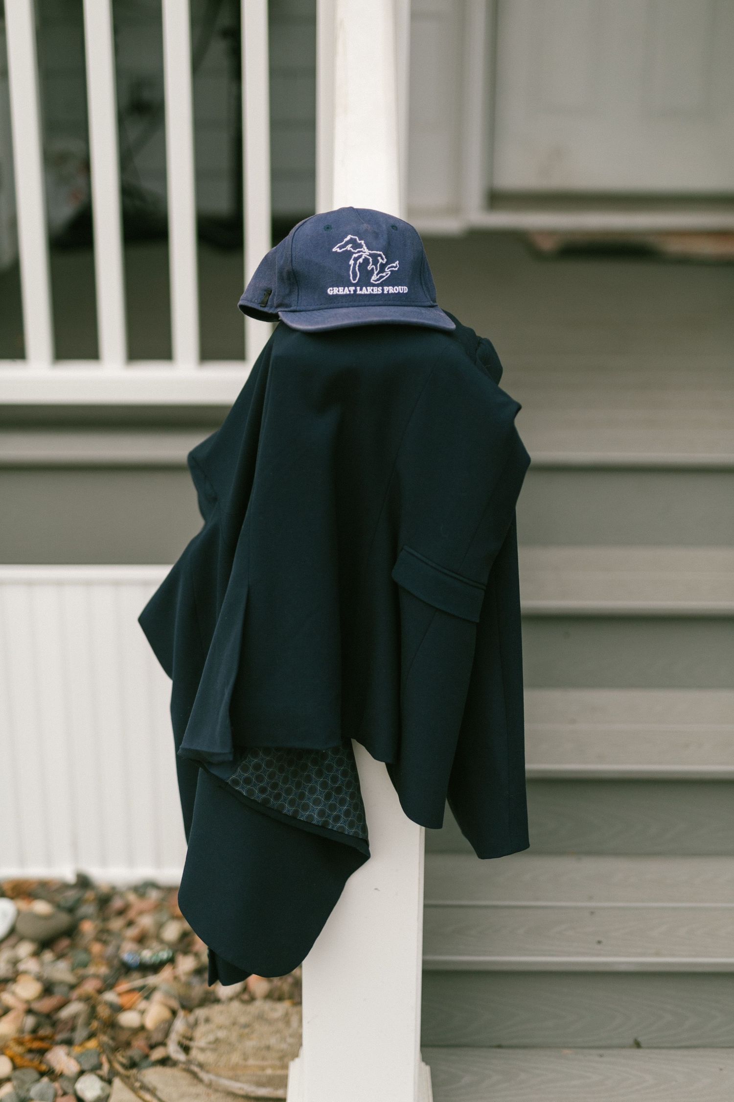 A navy blue baseball cap and black jacket draped over a white porch railing with stones visible below.