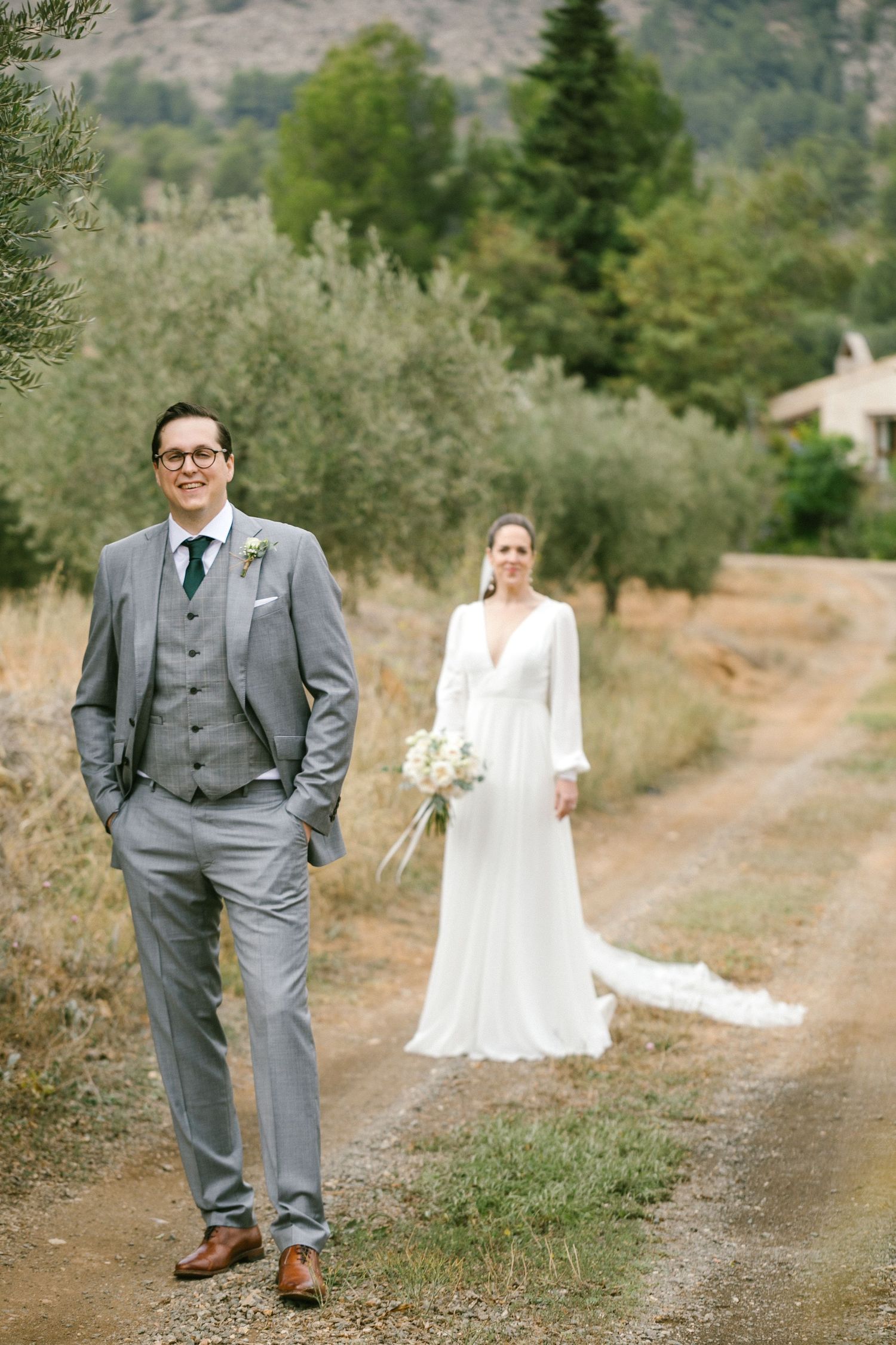 A bride and groom in wedding attire pose on a rustic dirt path with olive trees and greenery in the background.