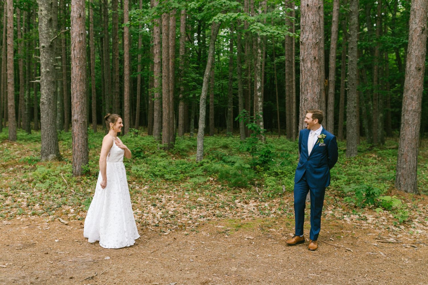 Two people in formal wedding attire stand apart in a serene pine forest setting.