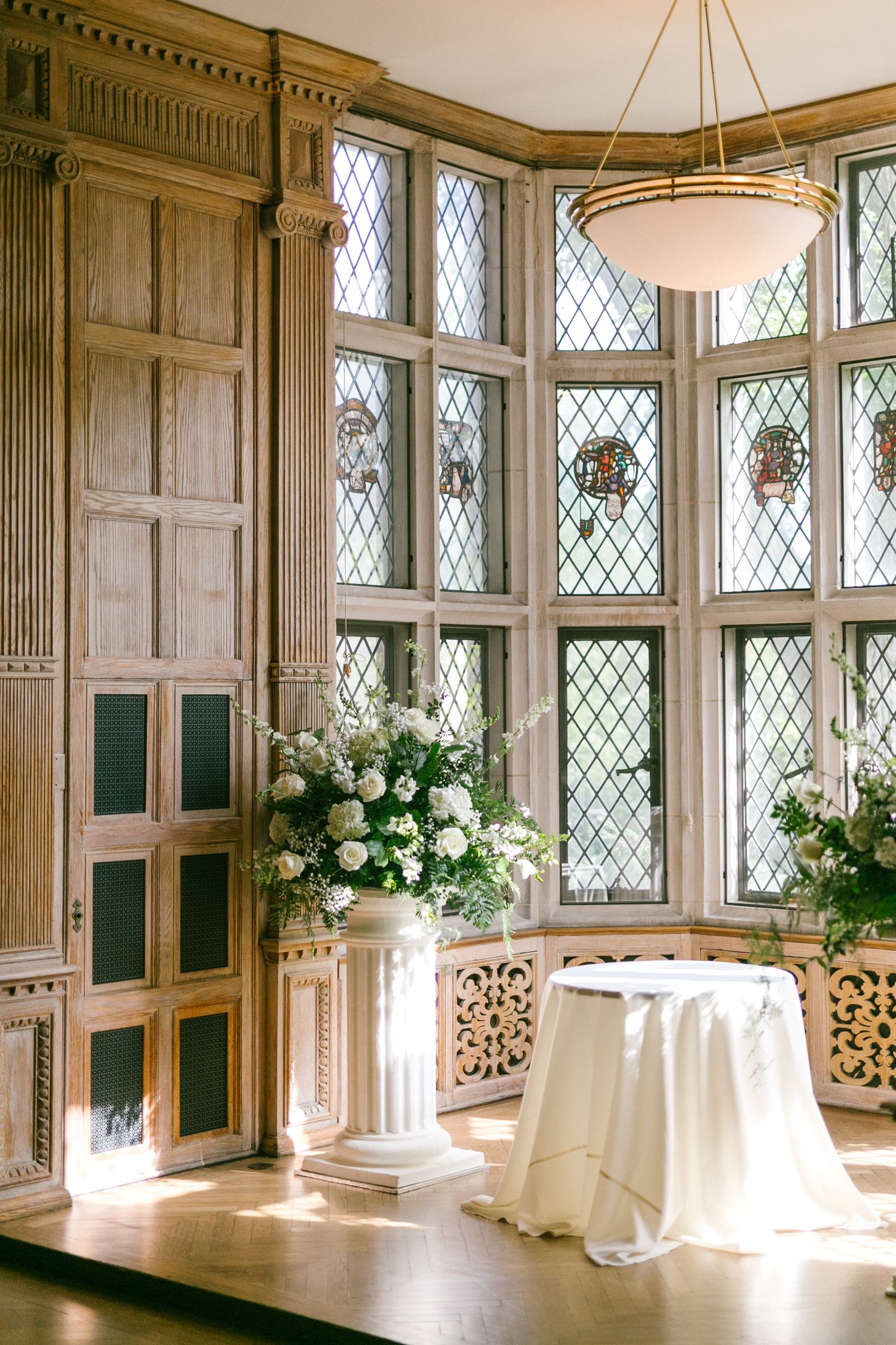 Elegant wedding venue with leaded glass windows, white floral arrangement on pedestal, and draped table in wood-paneled room.