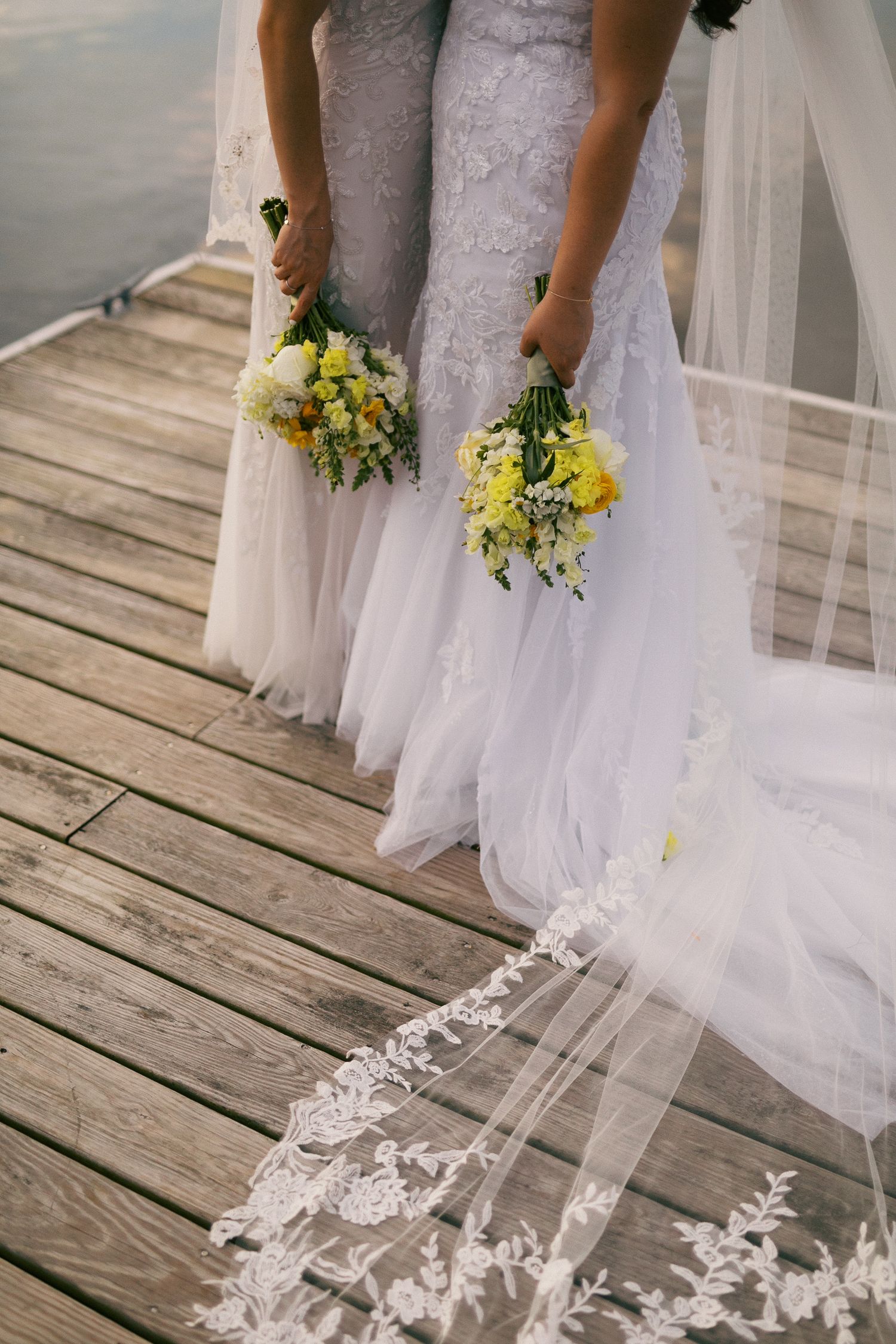 Two brides holding yellow flower bouquets stand on a wooden dock, one wearing a lace veil trailing on the ground.