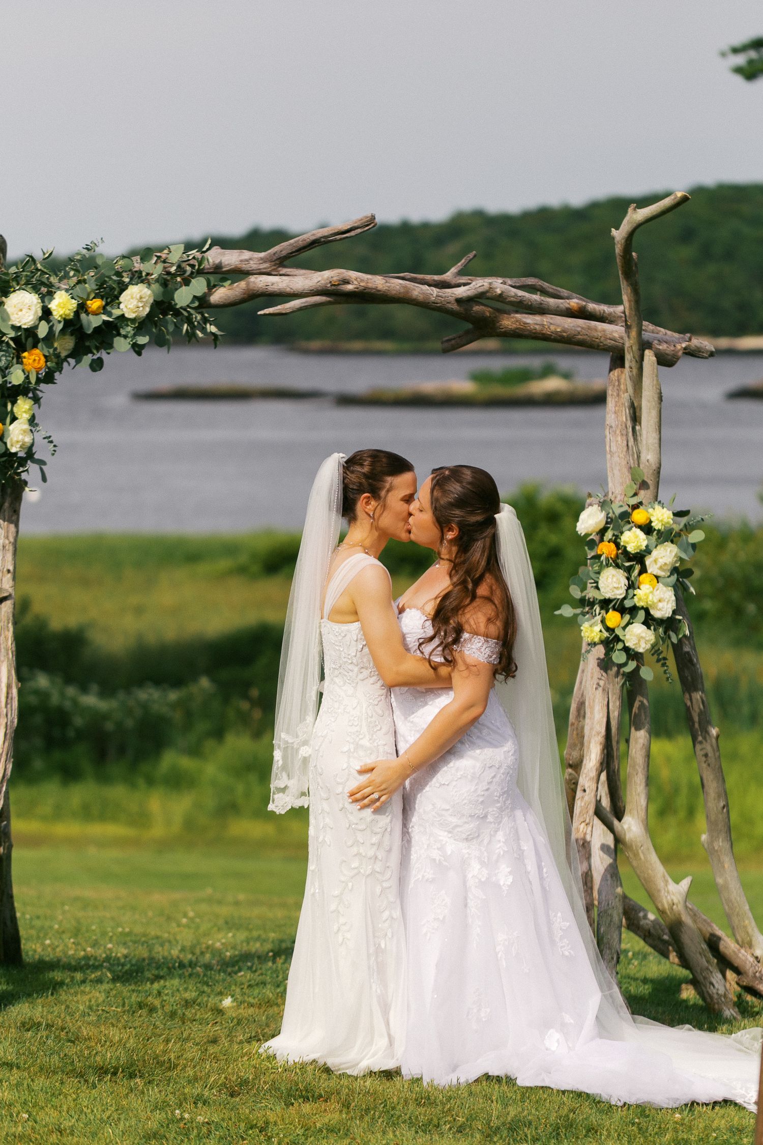 Two brides in white lace gowns share a kiss under a rustic driftwood arch adorned with yellow and white flowers by a waterfront.