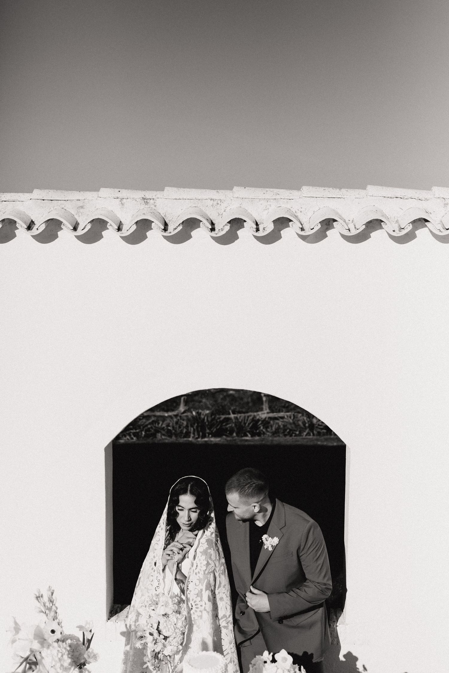 Black and white photo of bride and groom standing in an arched doorway of a white building.