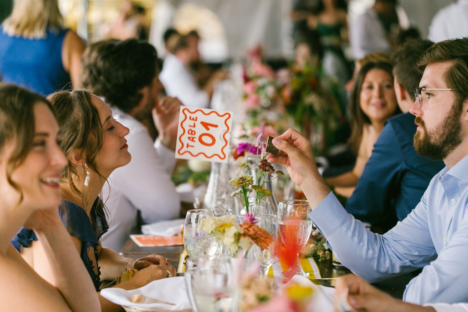 People enjoying a festive outdoor dining celebration with drinks and food at a long table in natural lighting.