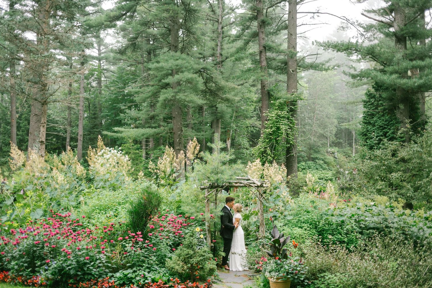 A garden wedding ceremony setting surrounded by evergreen trees and colorful flowers.