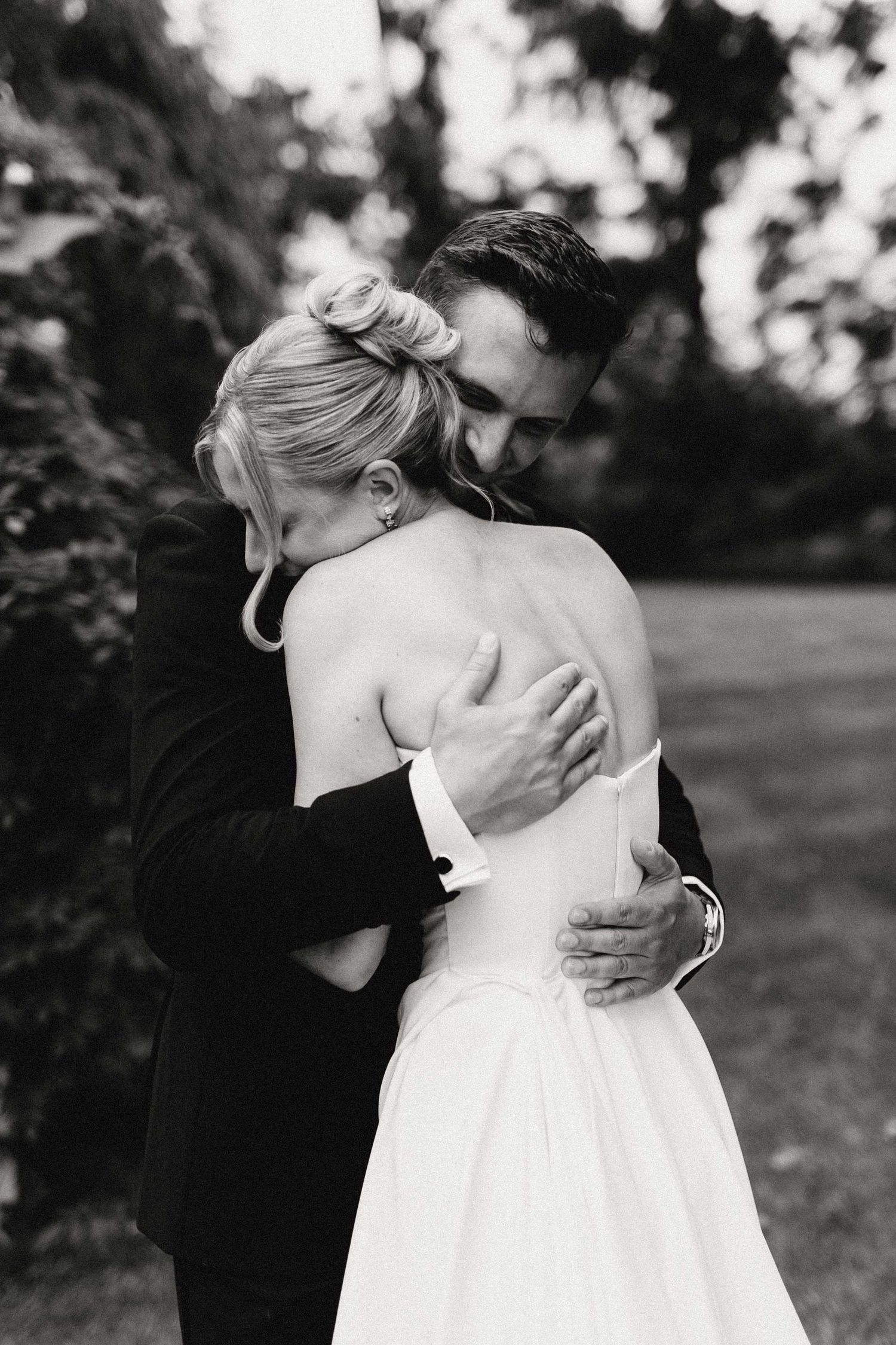 A tender black and white wedding photo capturing an intimate embrace between a couple in formal attire outdoors.