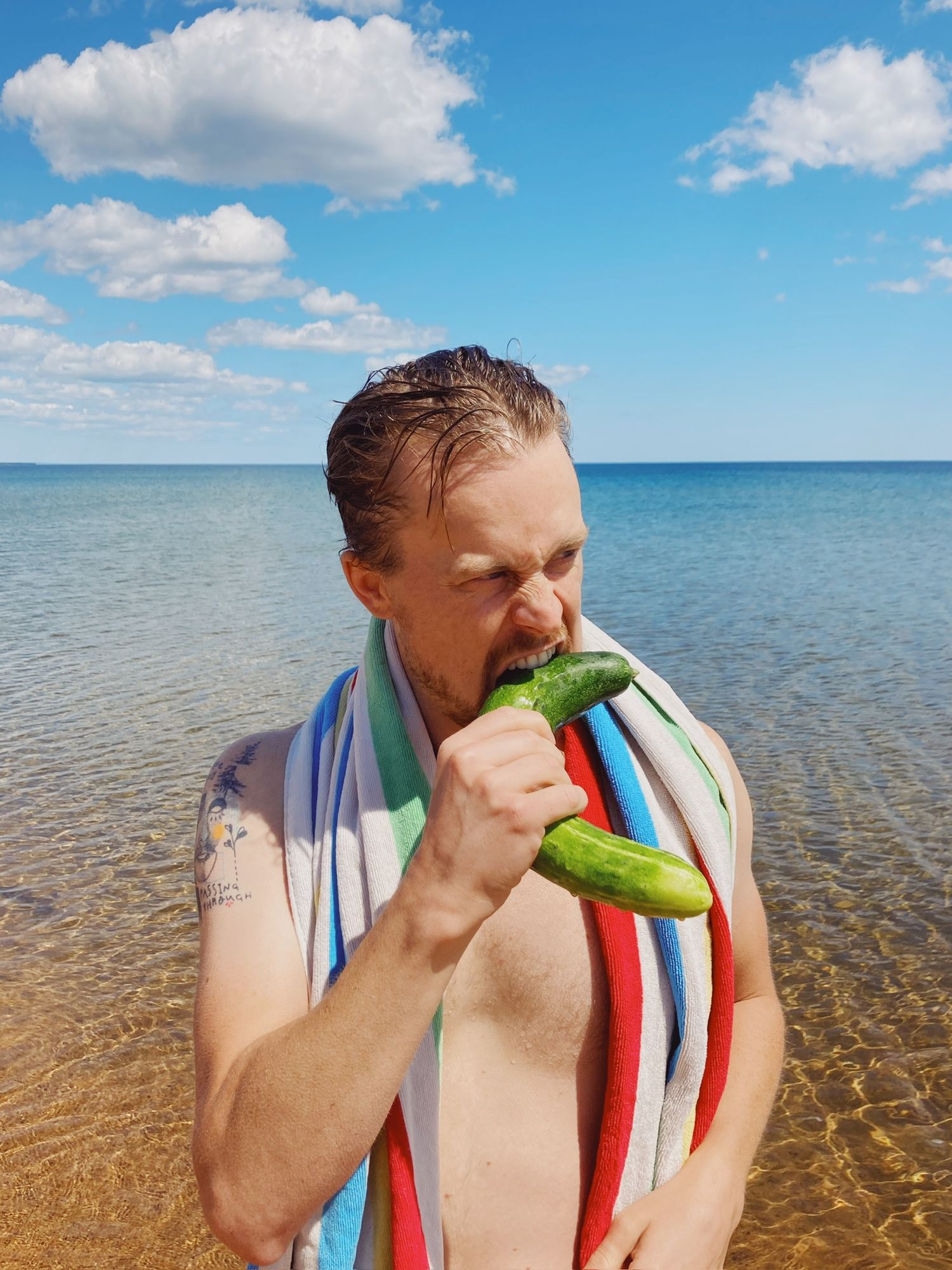 Shirtless man with a striped towel eating a cucumber on a sunny beach by the ocean.