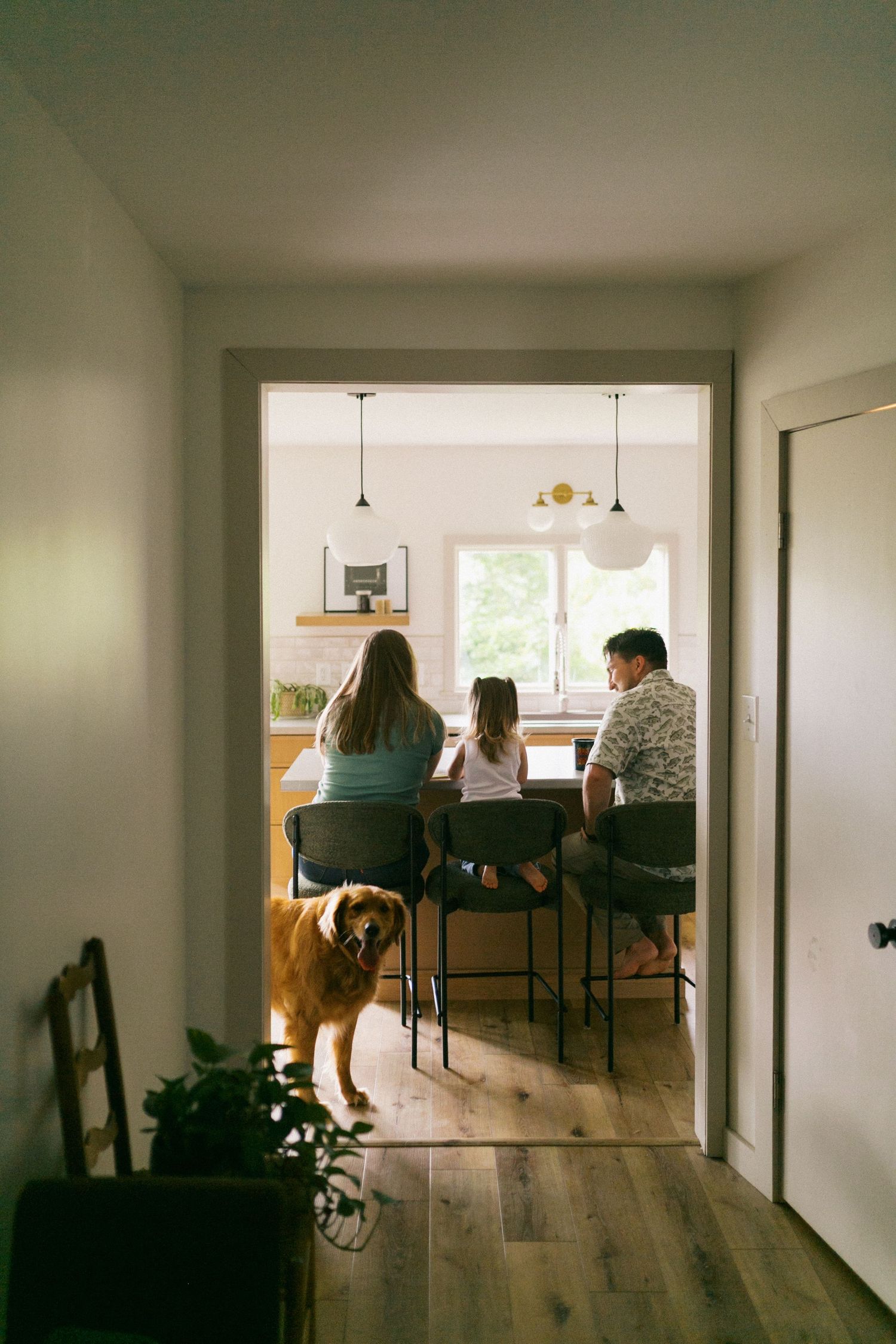 Family with golden retriever viewed from hallway, seated at kitchen counter facing window.