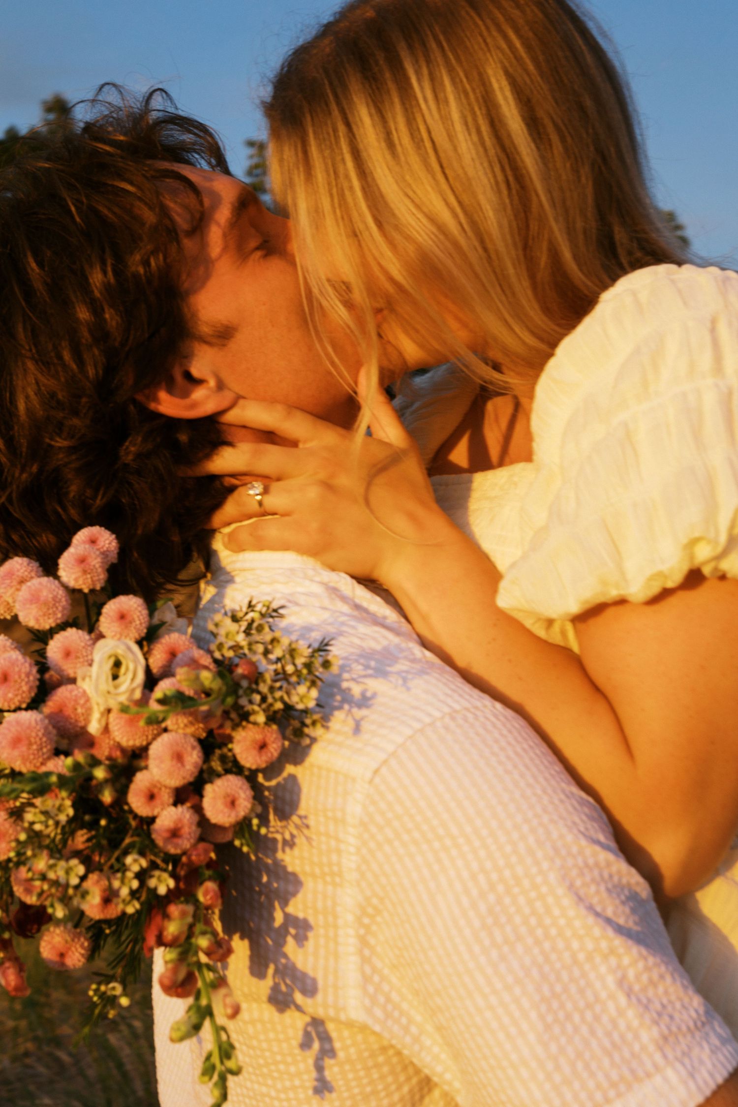 A couple shares a romantic kiss outdoors, with the man holding a bouquet of pink flowers, bathed in warm golden sunlight.