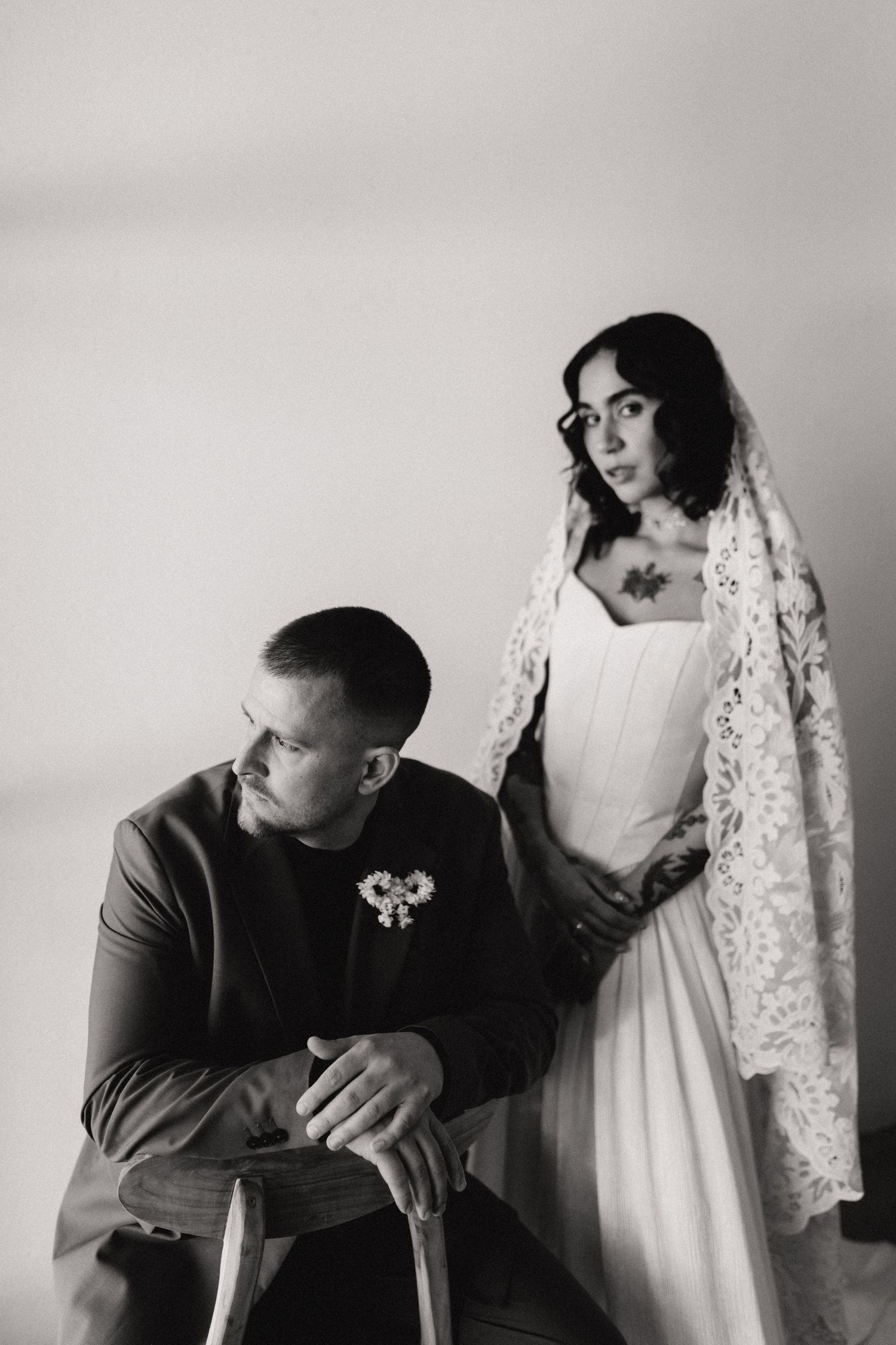 Black and white portrait of groom seated and tattooed bride in lace veil.