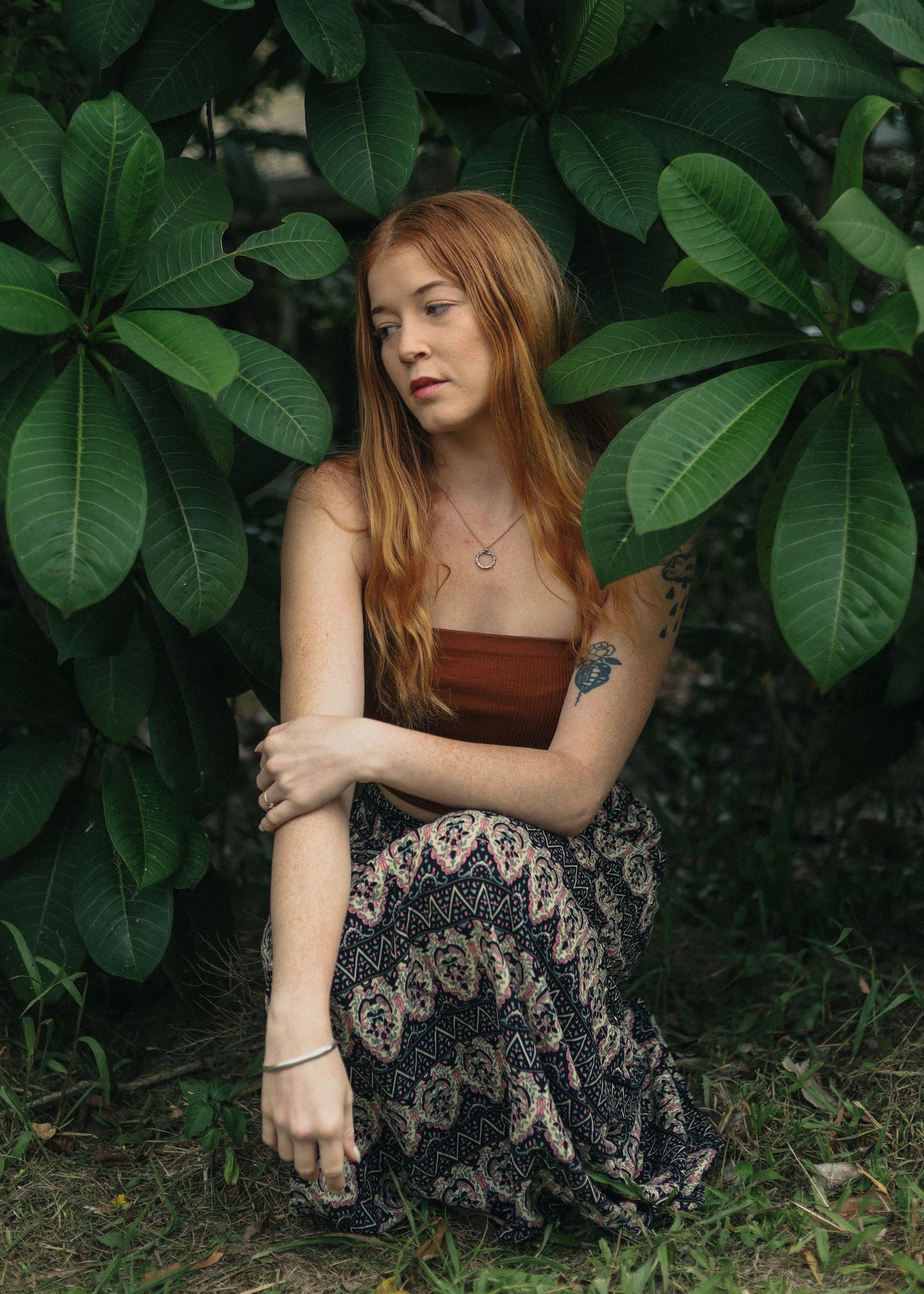 A young redhead woman with tattoos sits cross-legged in a boho maxi skirt among lush tropical leaves.