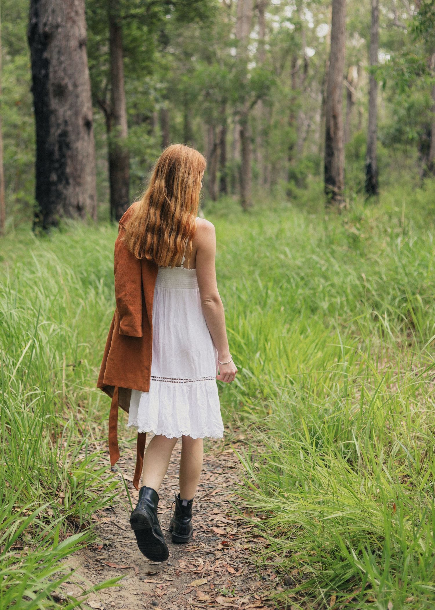 Young woman in white dress and brown jacket walks along a forest path surrounded by lush green trees.