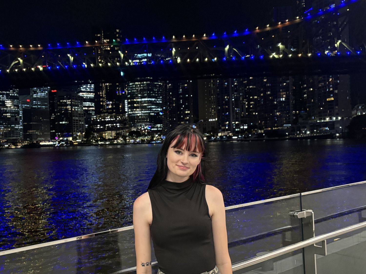 Woman in black dress poses by waterfront at night with illuminated bridge and city skyline backdrop.