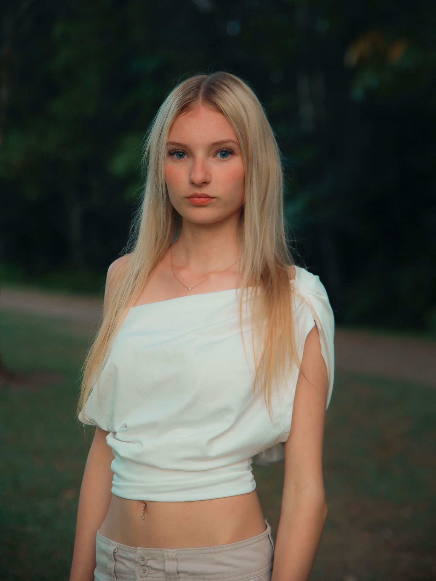 Young blonde woman in white off-shoulder crop top posing outdoors in a park at dusk.