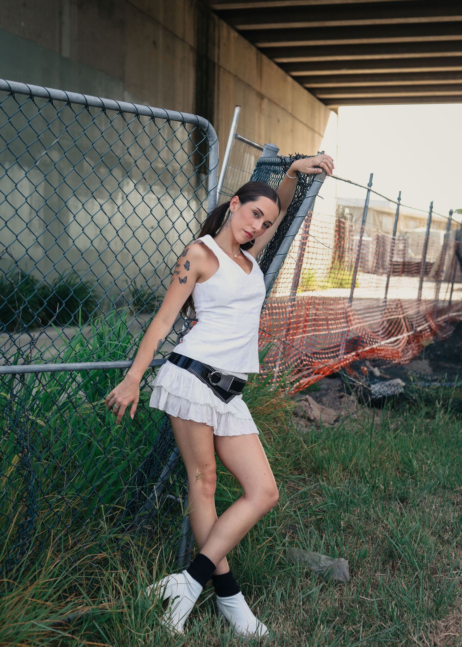 Young woman in white tennis skirt and tank top poses against chain-link fence in urban setting with orange safety netting.