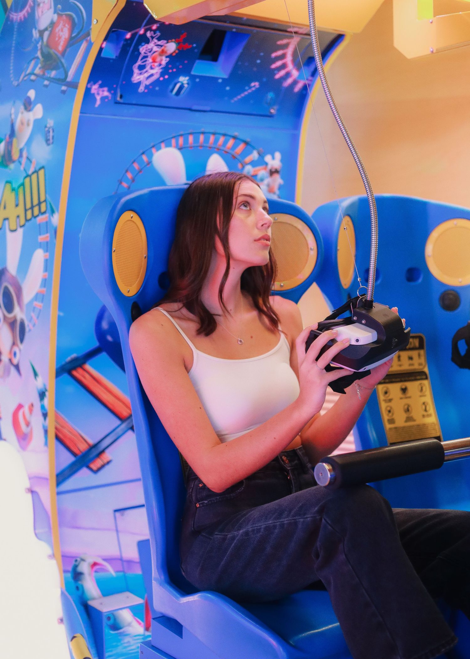 Young woman sitting in a colorful blue arcade game cabinet, holding a controller while looking up thoughtfully.