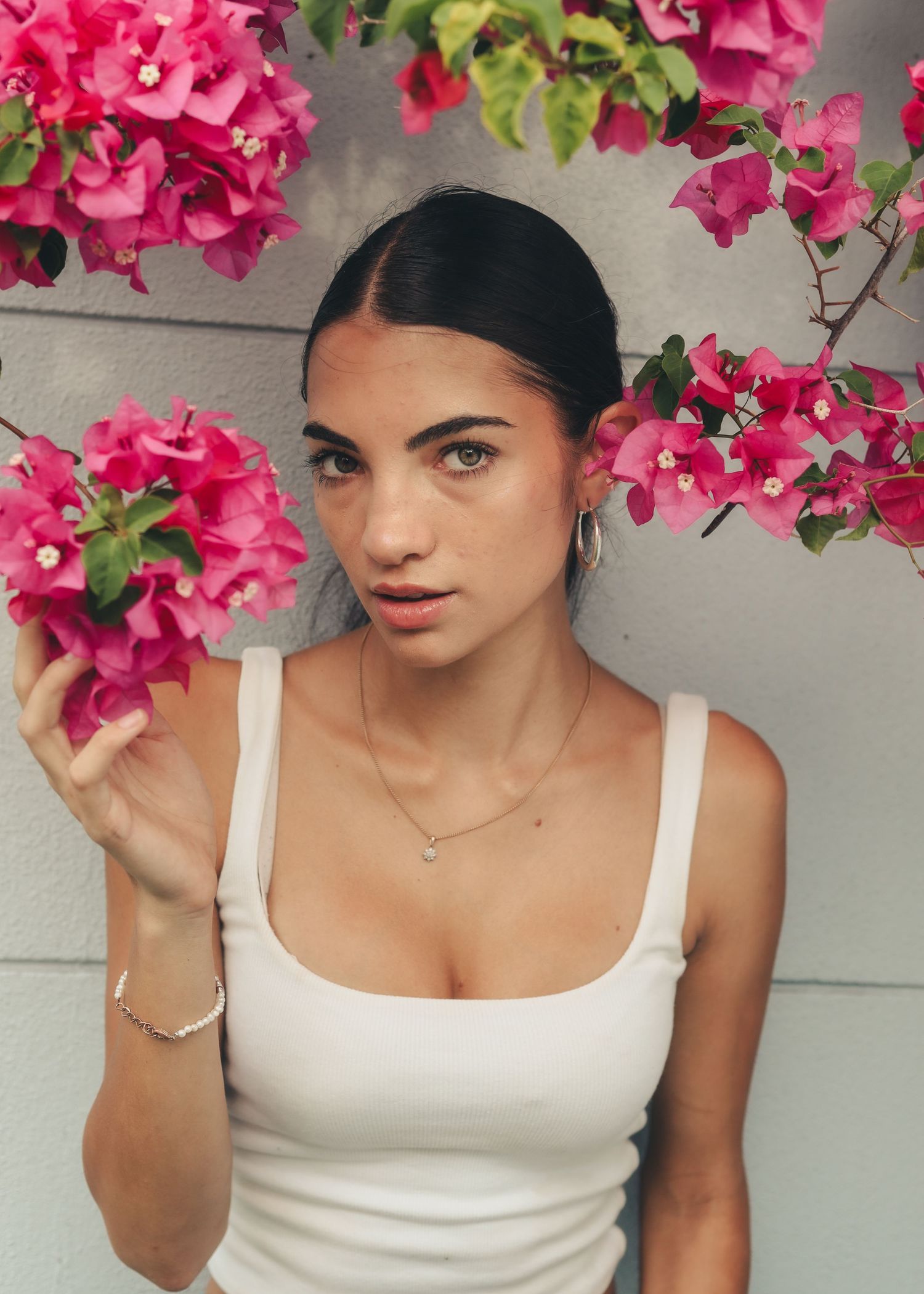 Young woman in white tank top holding pink bougainvillea flowers, wearing delicate jewelry, posing against a light wall.