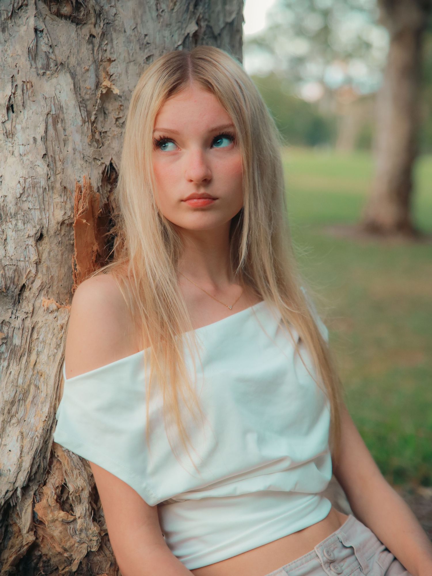 Young blonde woman in white off-shoulder top leaning against a tree in a park, gazing upward thoughtfully.