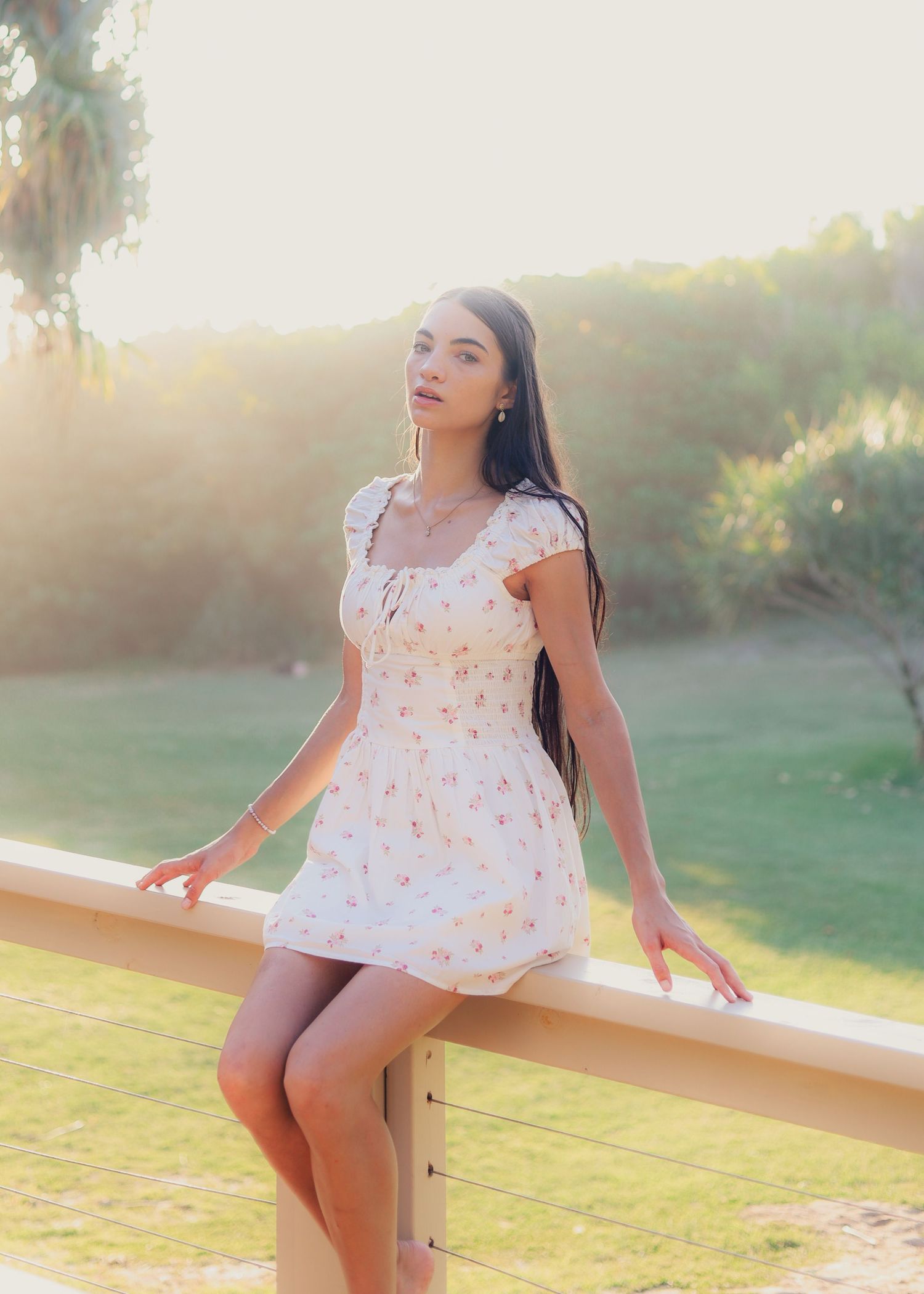Young woman in floral mini dress posing by a railing in golden hour sunlight with lush greenery background.