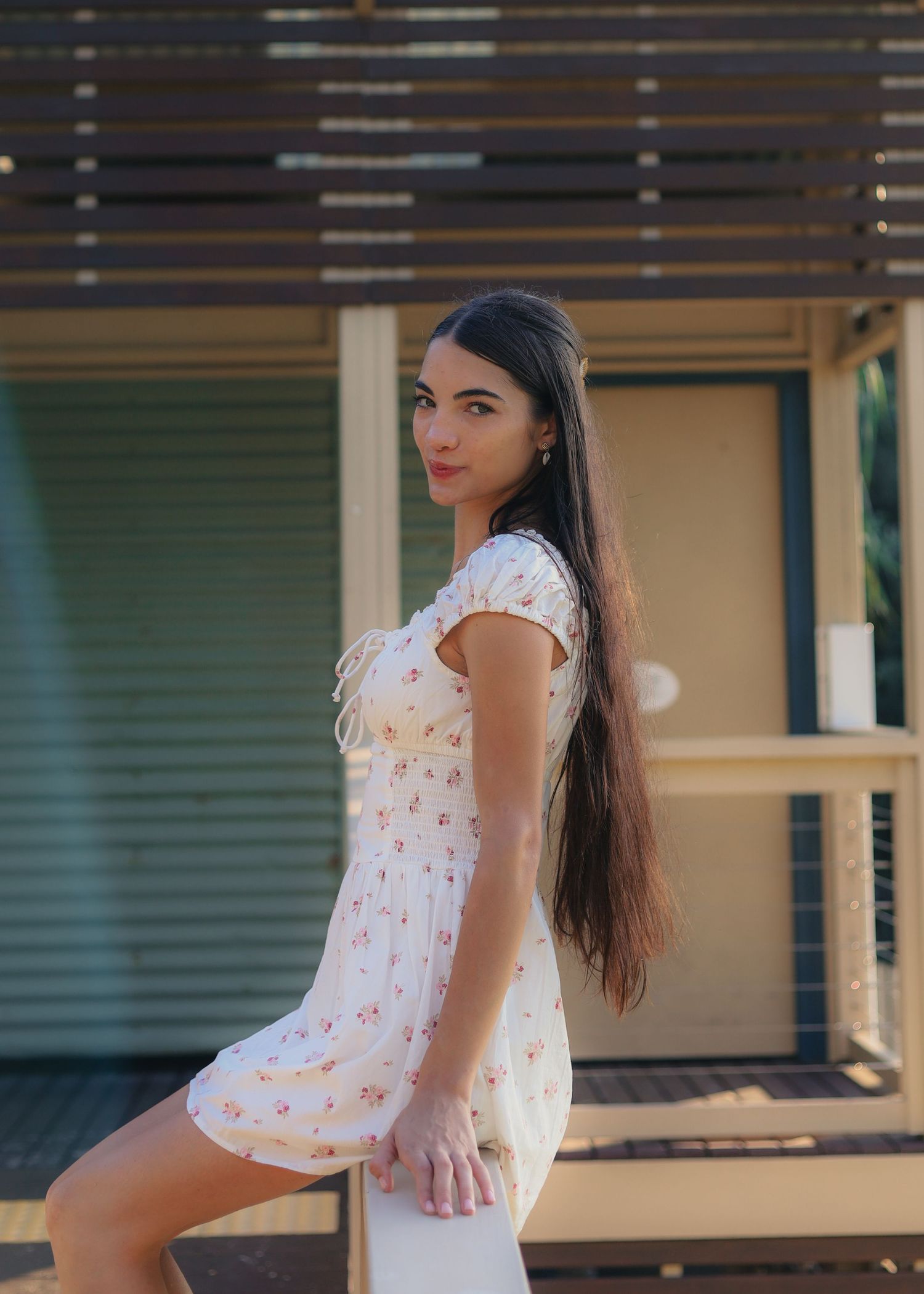 Young woman in floral mini dress posing outdoors near wooden structure on sunny day.