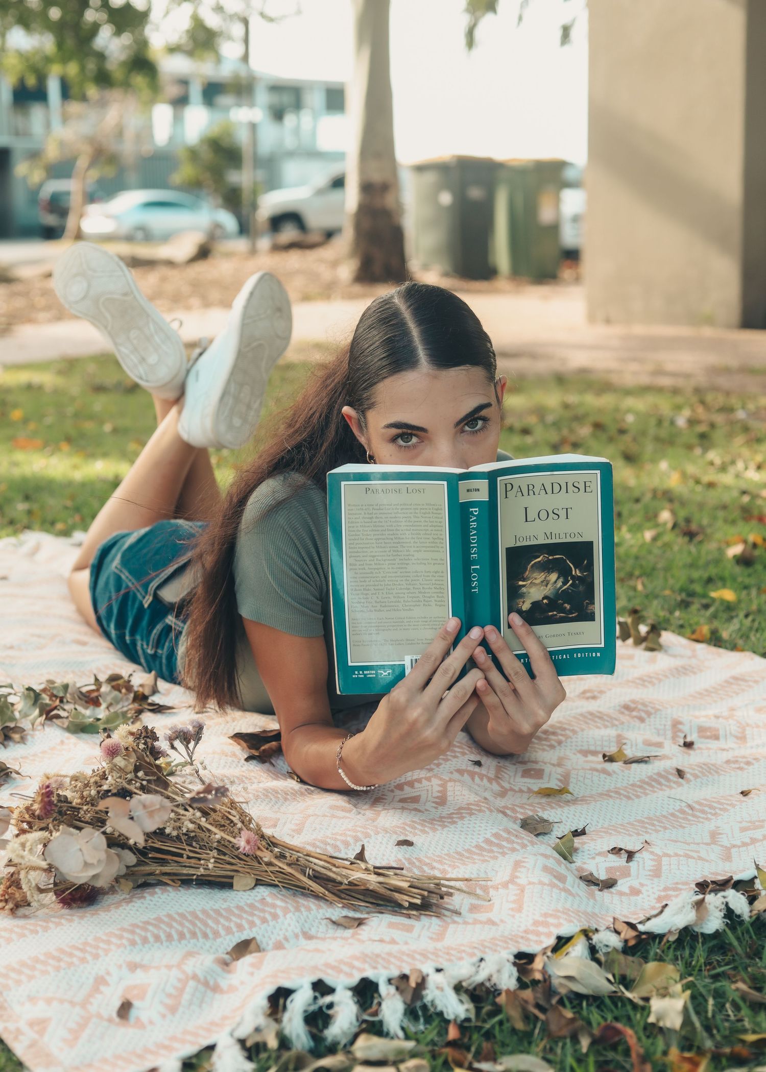 Young woman lying on a blanket outdoors reading Paradise Lost book, with flowers nearby and trees in background.