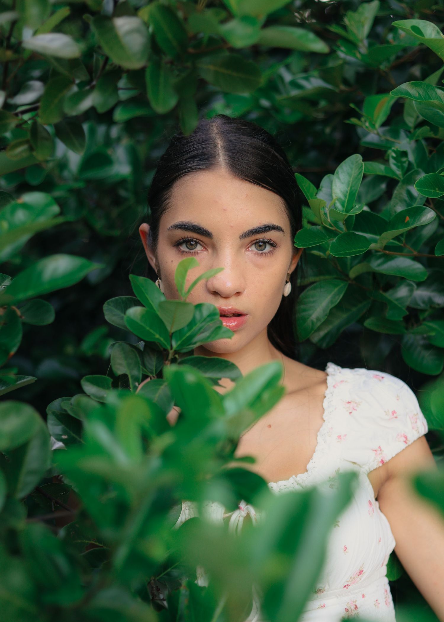 Young woman with dark hair in white floral dress peers through lush green tropical leaves in a garden setting.