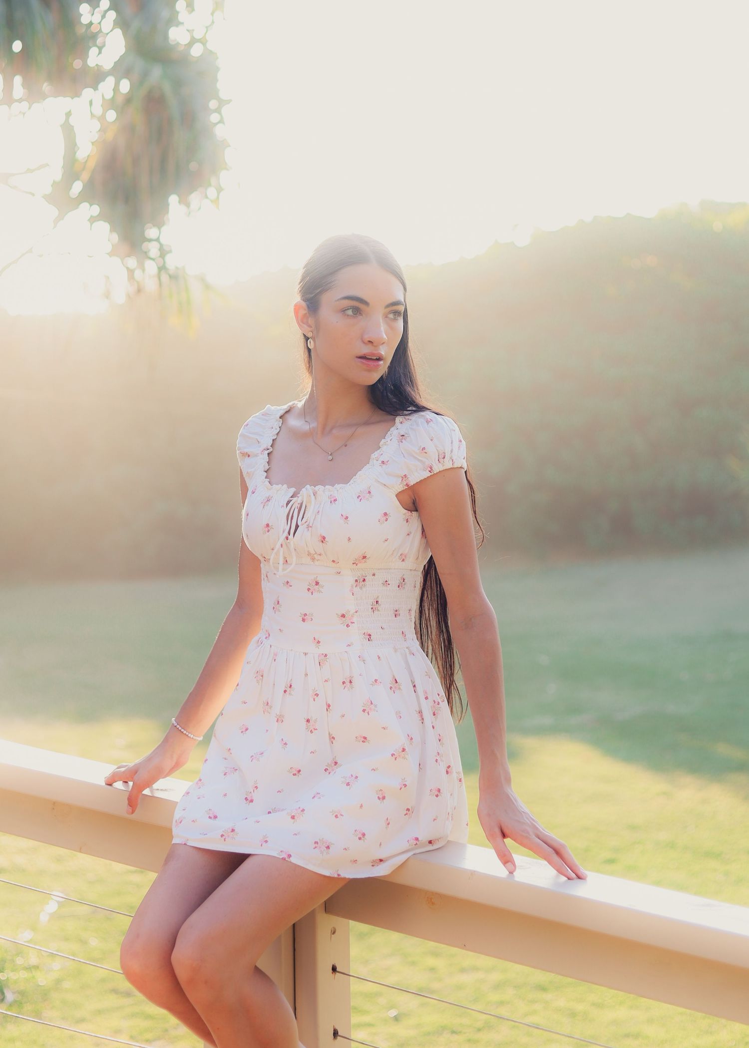 Woman in white floral mini dress posing by a wooden fence at golden hour with green hills behind.