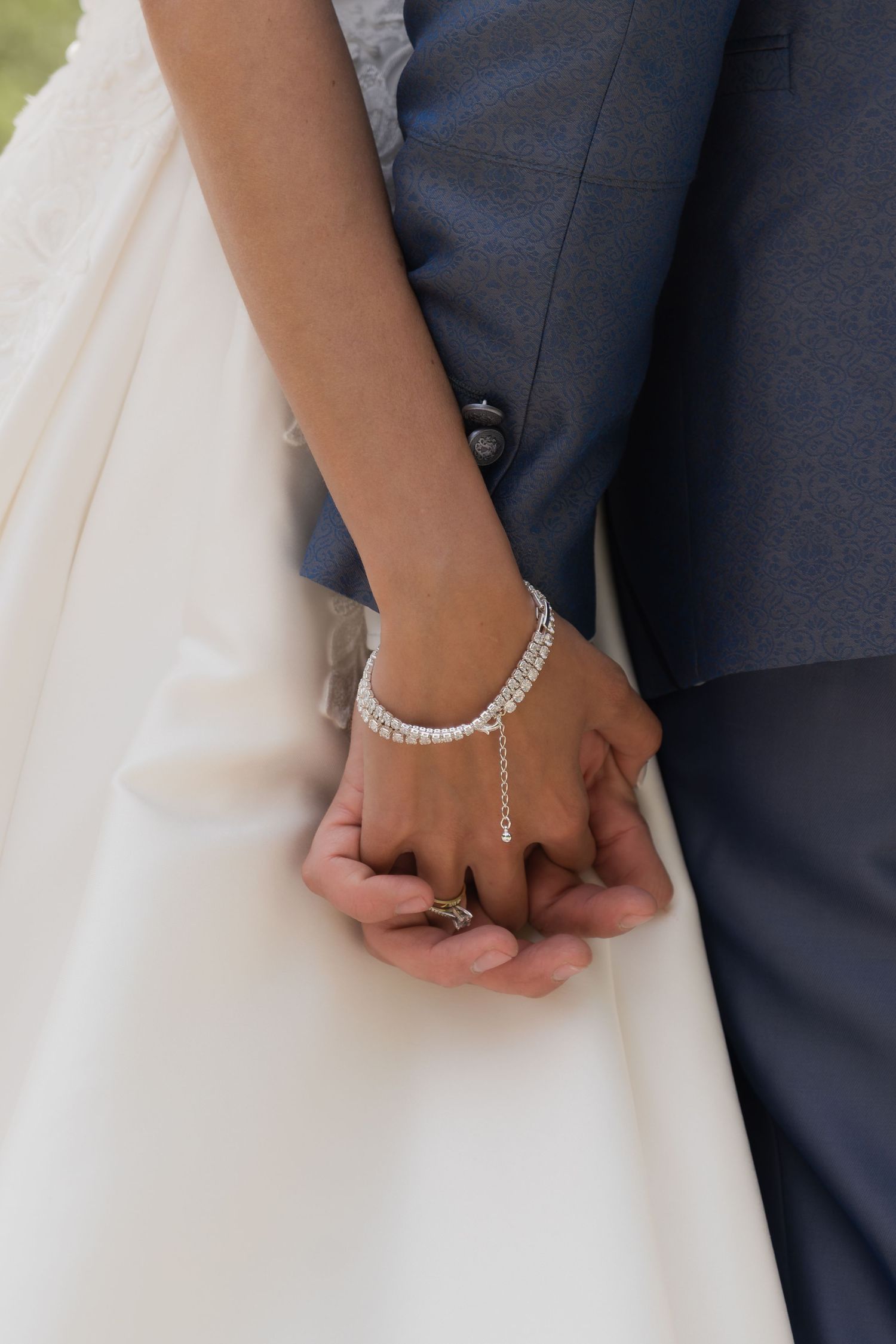 Close-up of bride and groom holding hands, bride wearing crystal bracelet and groom wearing gold wedding ring.