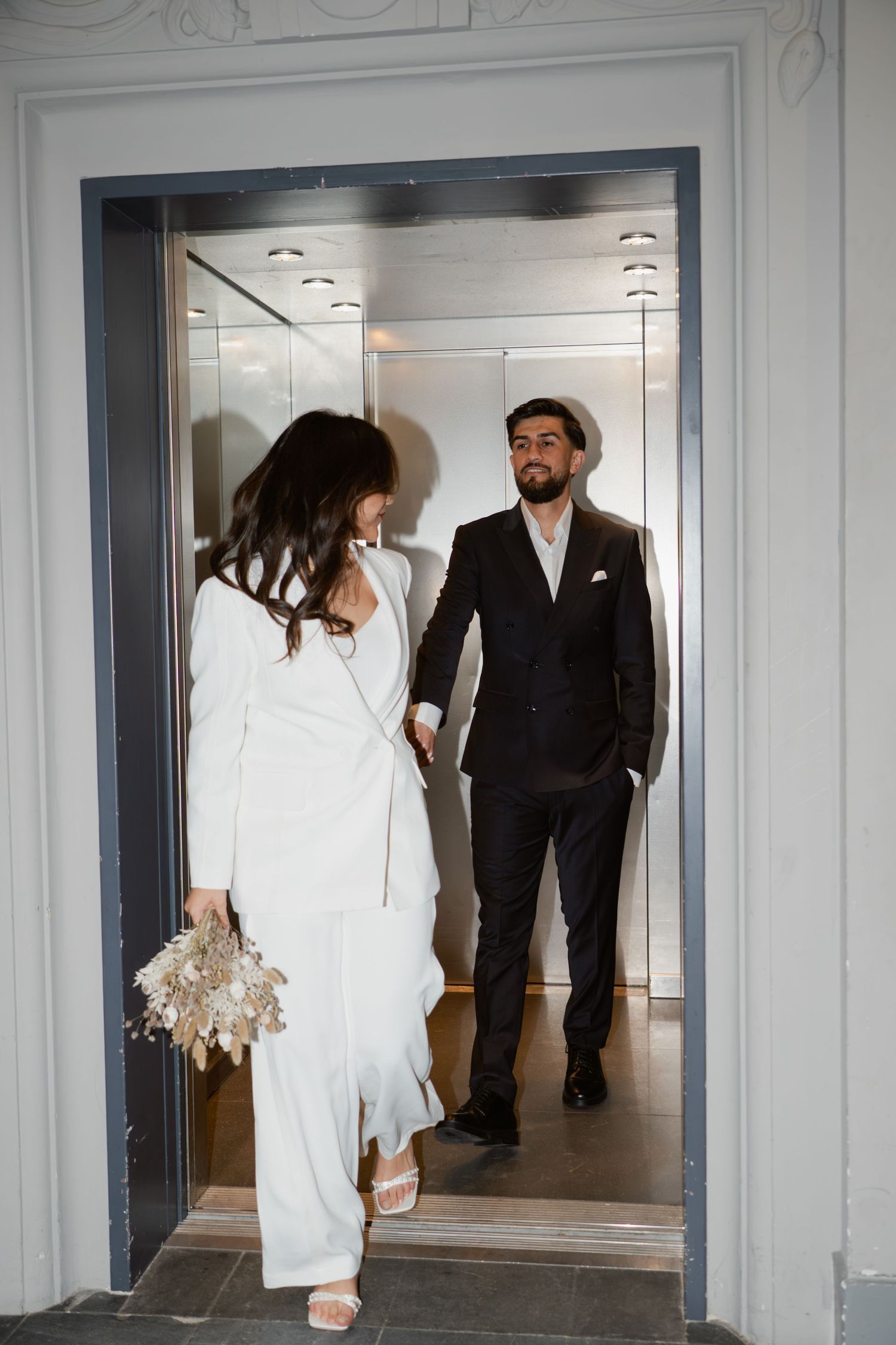 Bride in white pantsuit holding dried bouquet exits elevator with groom in black suit.