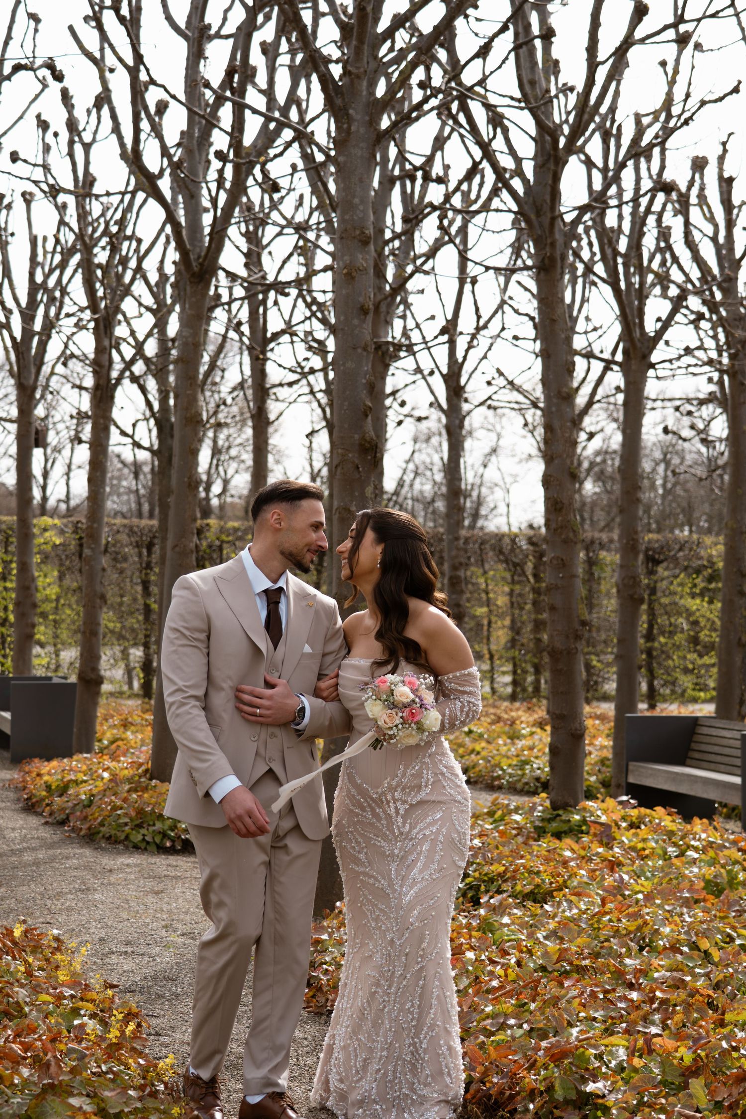 Wedding couple in autumn park, bride in beaded gown holding bouquet, groom in tan suit gazing lovingly.