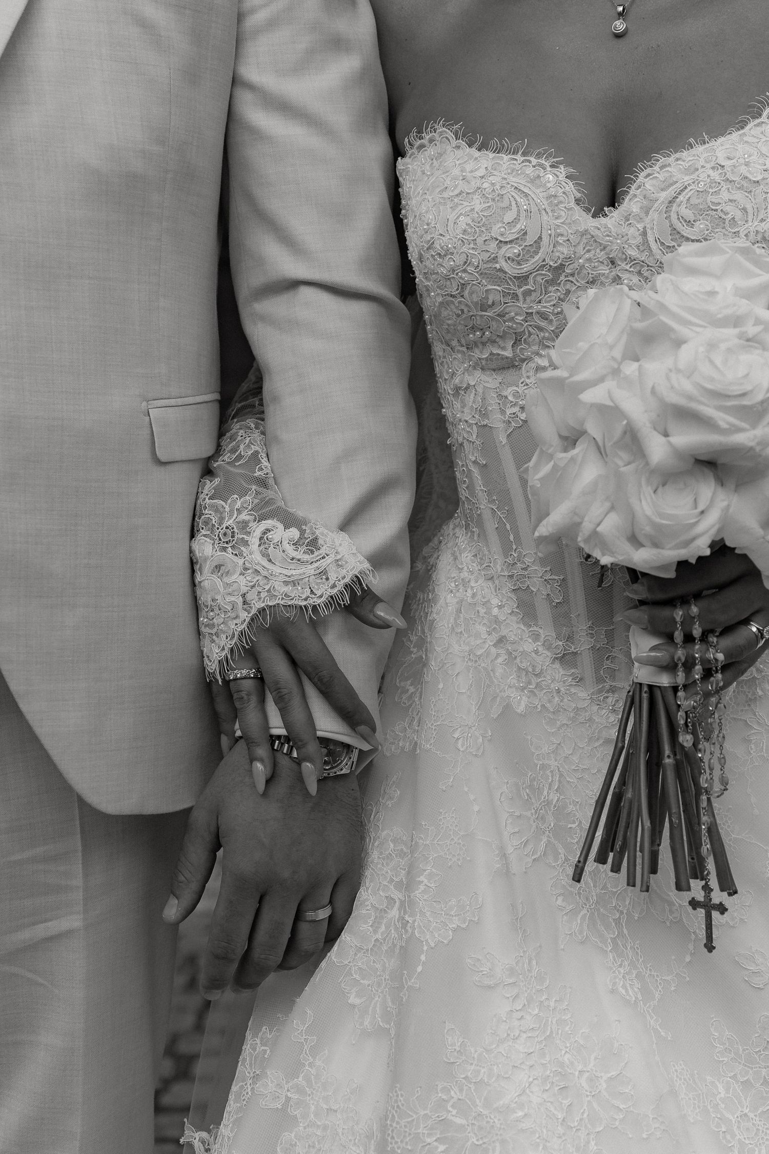 Black and white wedding photo of bride and groom holding hands, bride holding rose bouquet with cross, wearing lace dress.