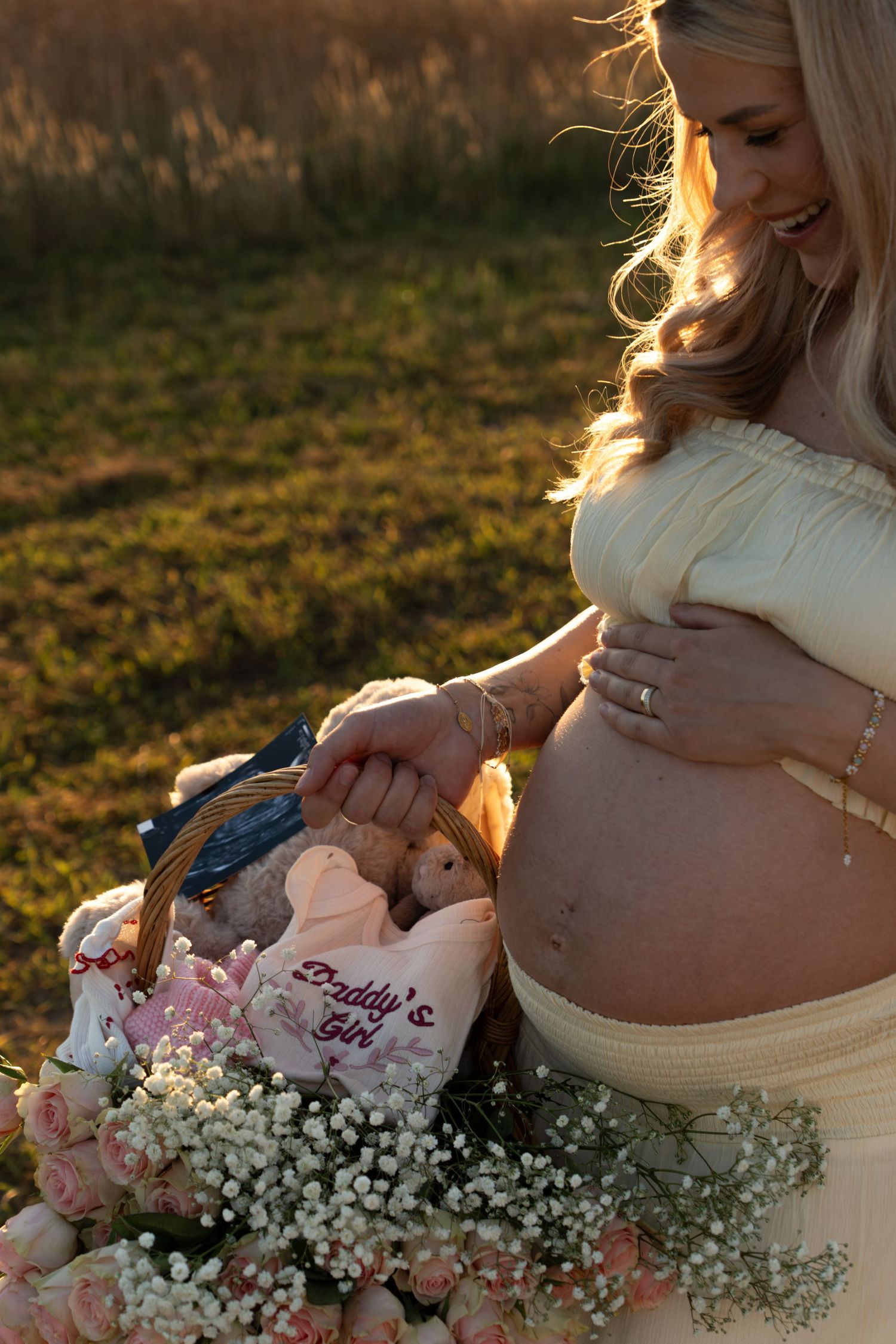 Pregnant woman sitting with floral basket containing Daddy's Girl onesie at sunset.