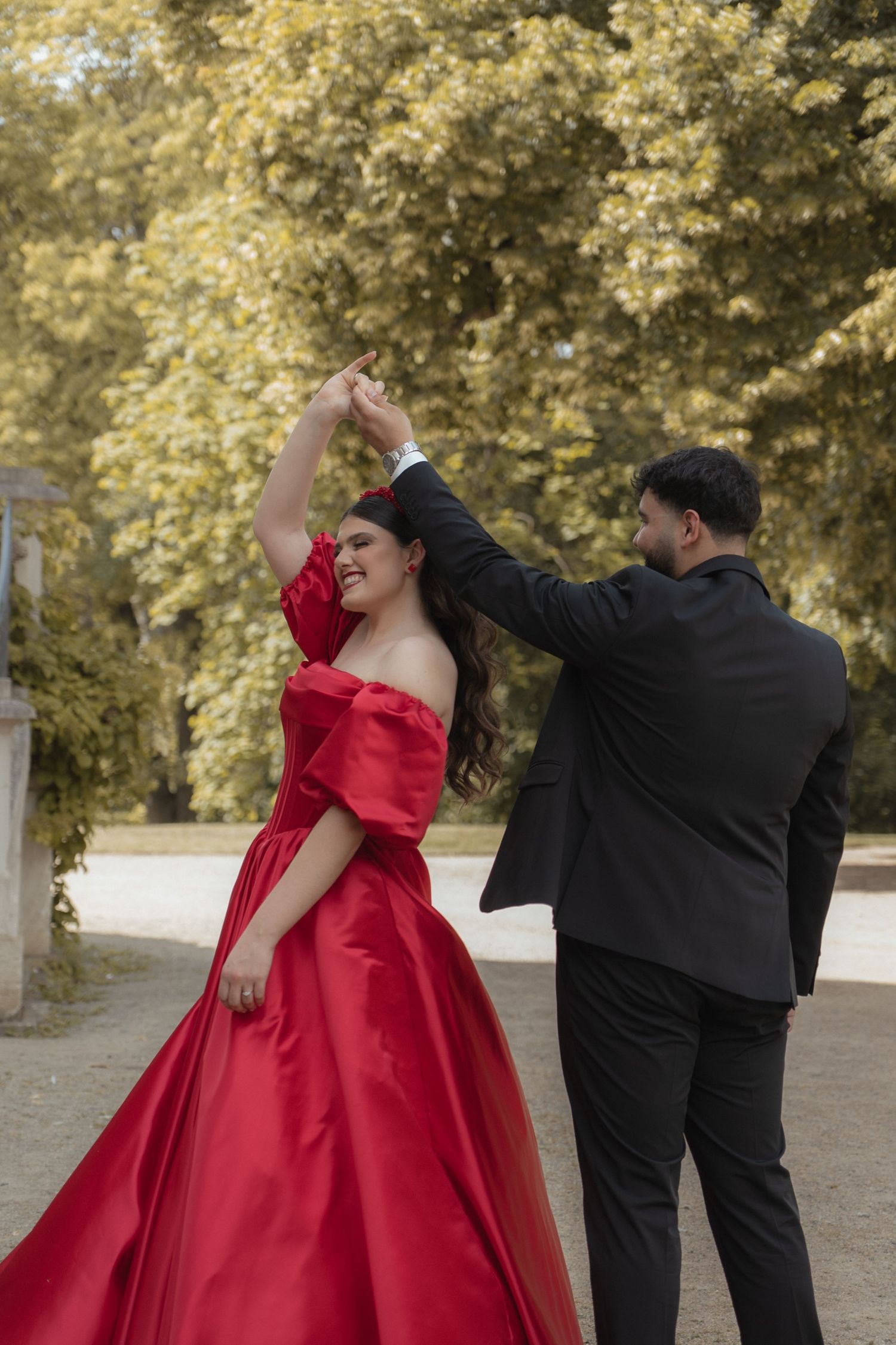 Couple dancing outdoors, woman in elegant red off-shoulder ball gown twirling with man in black suit.