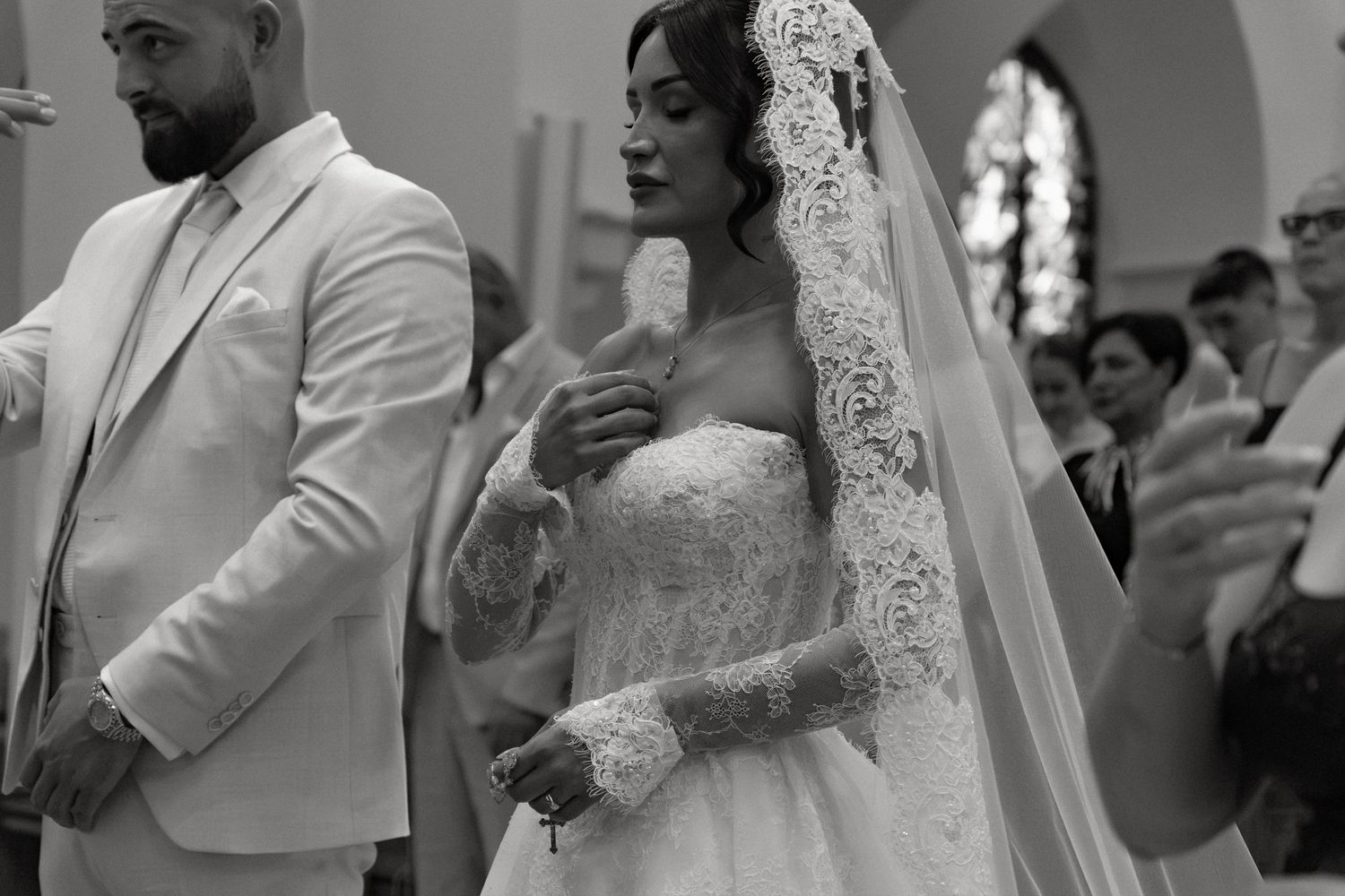 Black and white wedding photo of bride in lace gown and veil holding mic, groom in suit beside her in church.