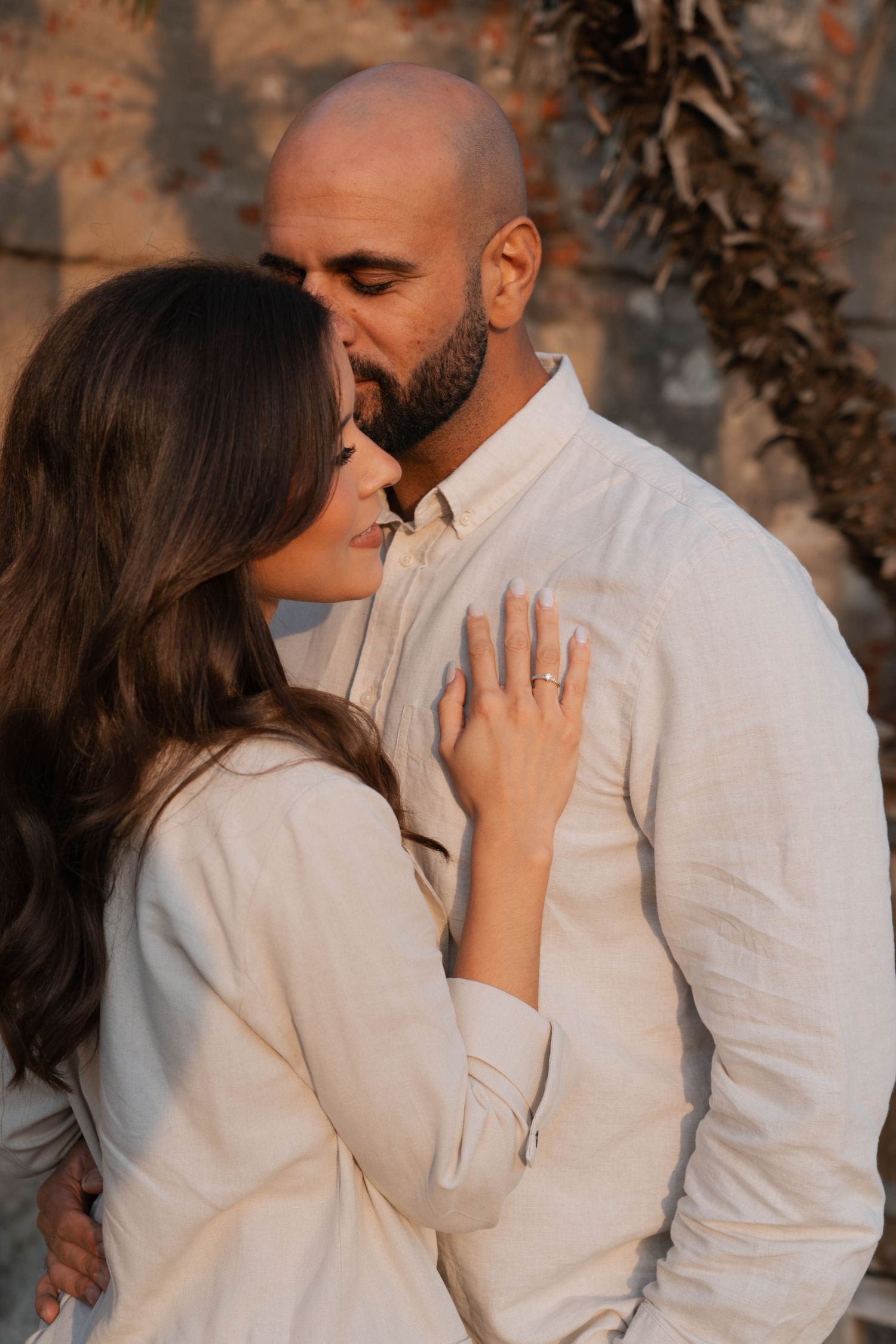 Bald man kissing woman with engagement ring against stone wall at golden hour.