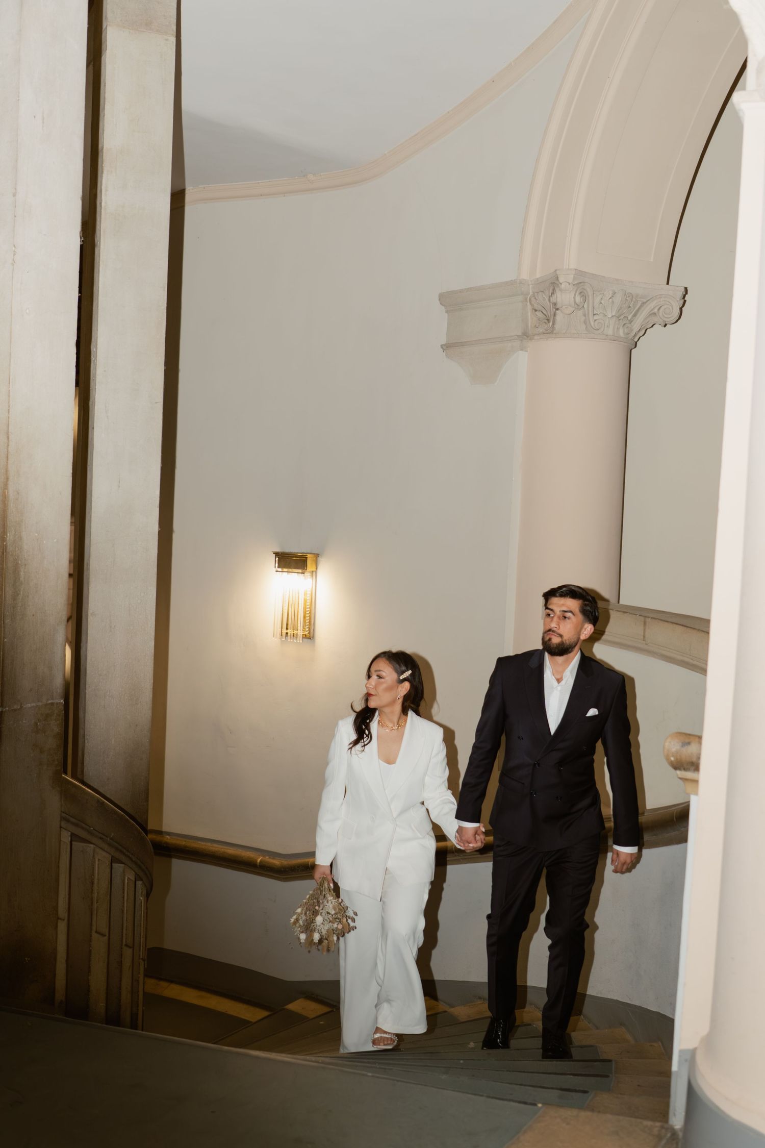 Bride and groom in white and black formal wear holding hands walking up elegant staircase.
