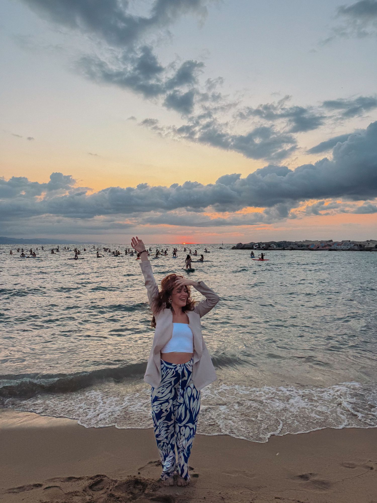Woman in blue floral pants and white crop top posing on a beach at sunset with arm raised.