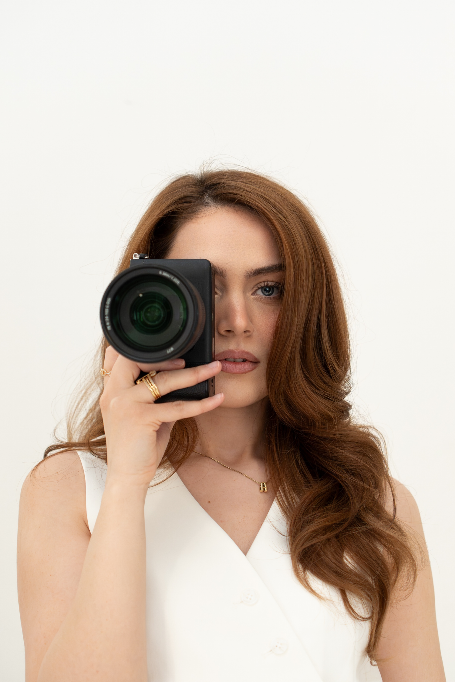 Young woman with auburn hair holding camera in front of face, white background studio shot.