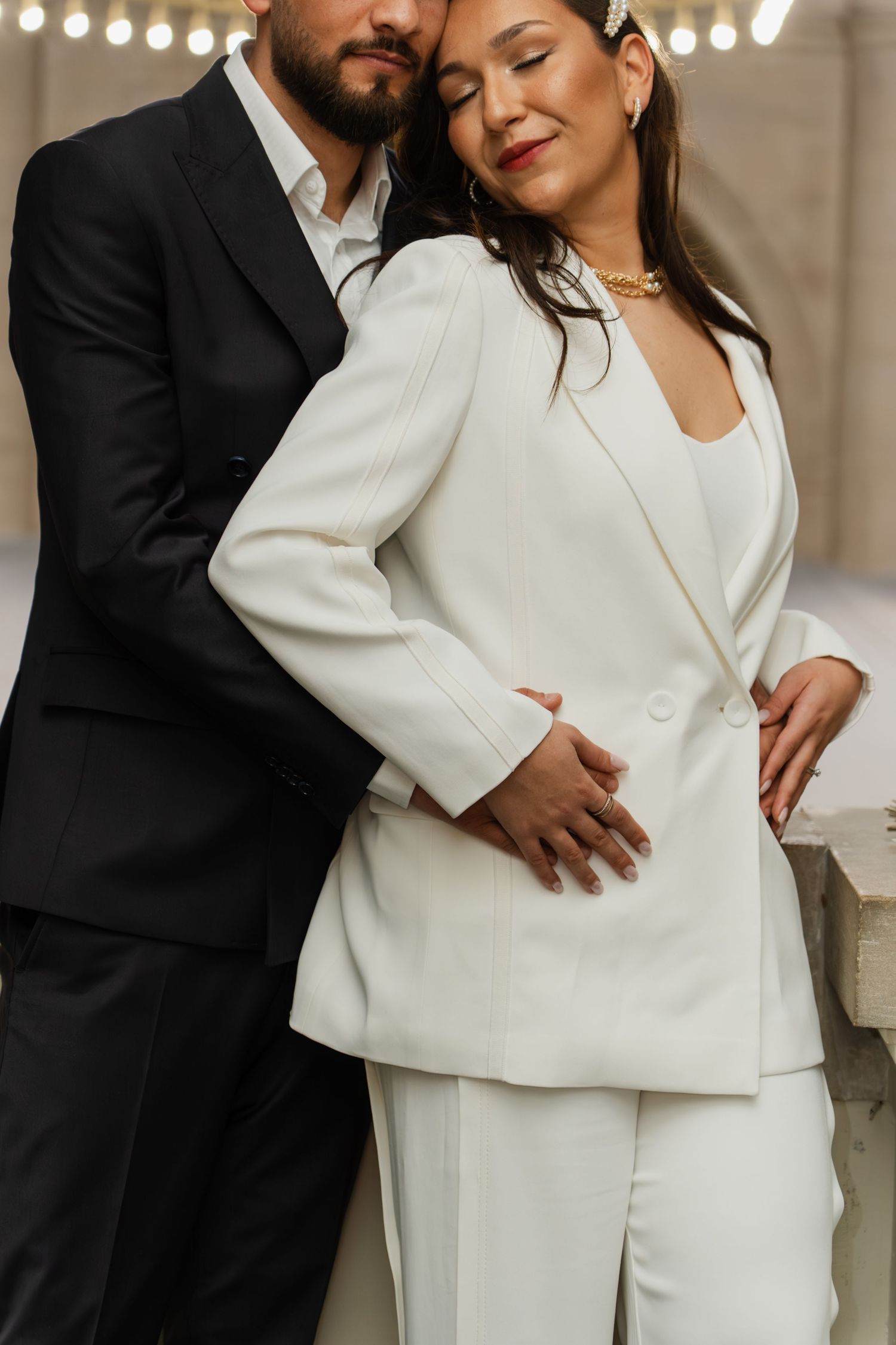 Elegant couple embracing, woman in white blazer suit and man in black tuxedo at a romantic indoor setting.
