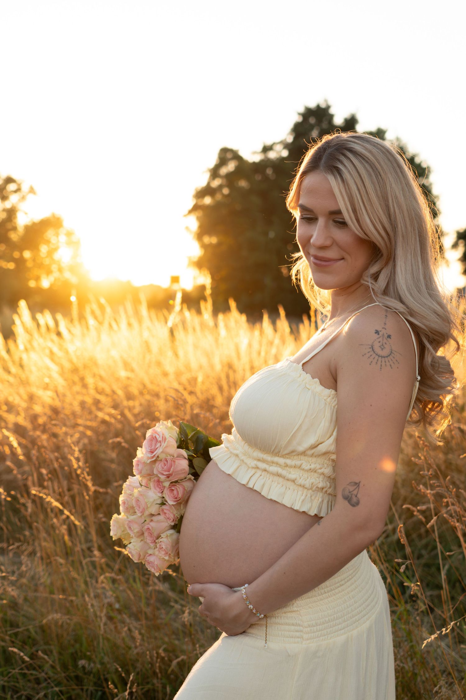 Pregnant blonde woman in yellow dress holding pink roses bouquet at golden hour sunset.