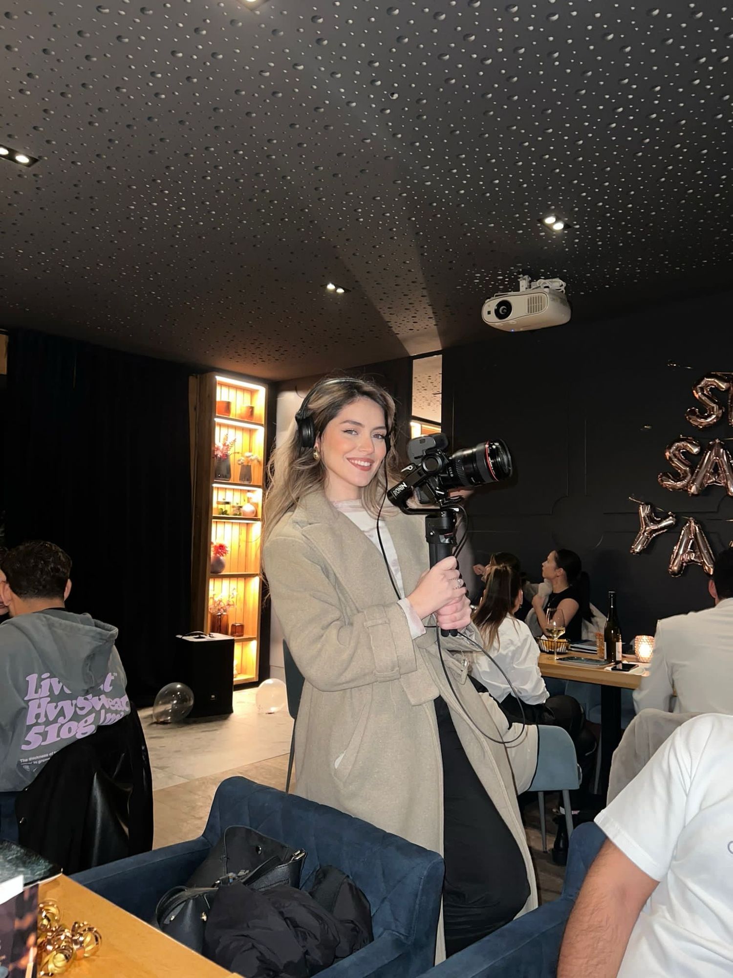 Smiling blonde woman holding a professional gimbal camera in a modern dark-ceiling restaurant or event venue.
